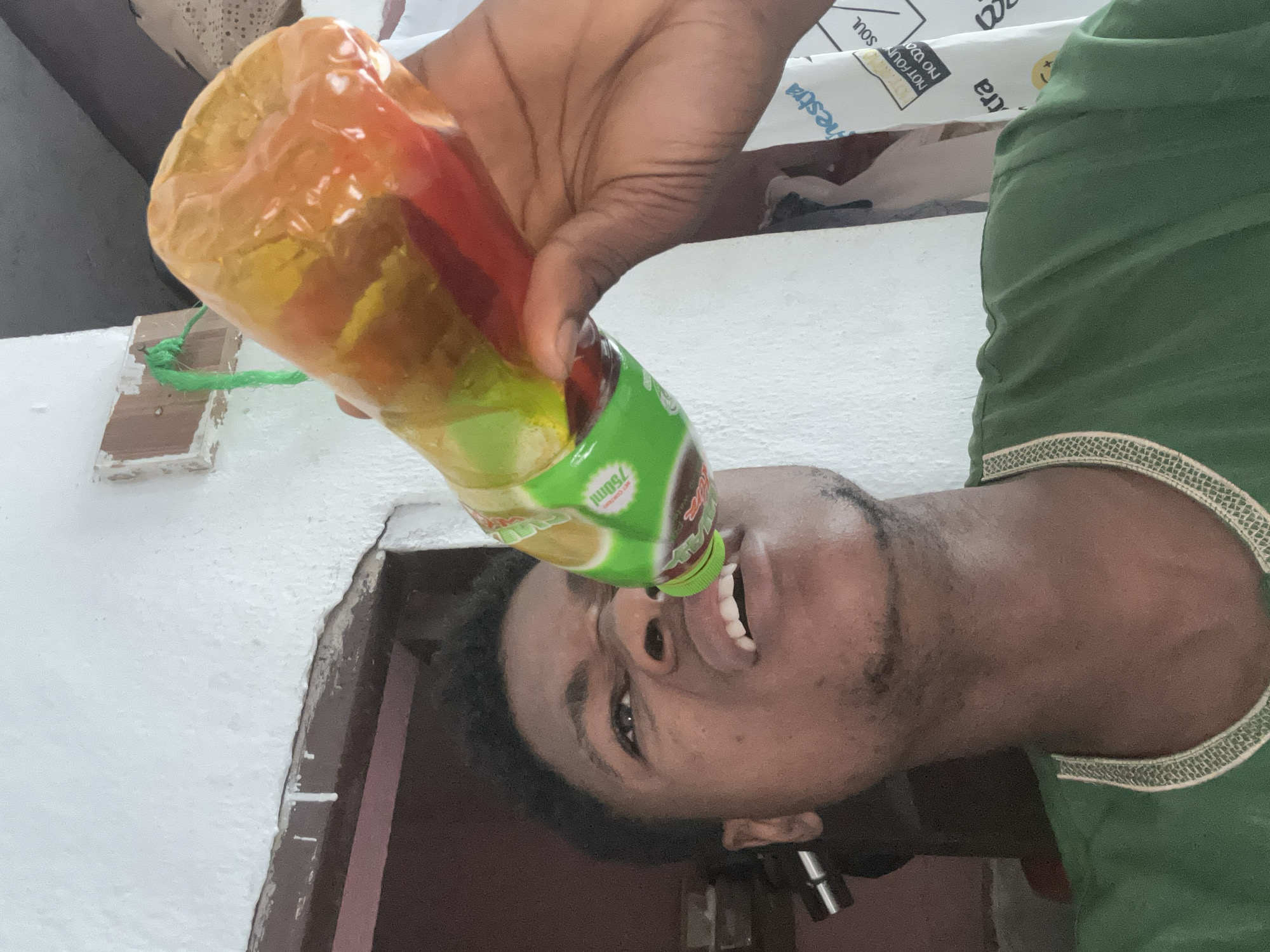 A close-up, low-angle shot captures a young Black male in Ihiagwa, Nigeria, drinking from a plastic bottle of liquid. The man is wearing a green garment with decorative trim around the neckline. His eyes are wide open, and his mouth is slightly ajar as he tilts the bottle to drink. The bottle, filled with a multi-colored liquid, is held in his right hand. Behind him, a white-painted wall with some peeling paint is visible, along with a dark doorway and a piece of cloth hanging on the wall with text. A green string is attached to a piece of wood on the wall. The lighting suggests an indoor setting, possibly during the day.