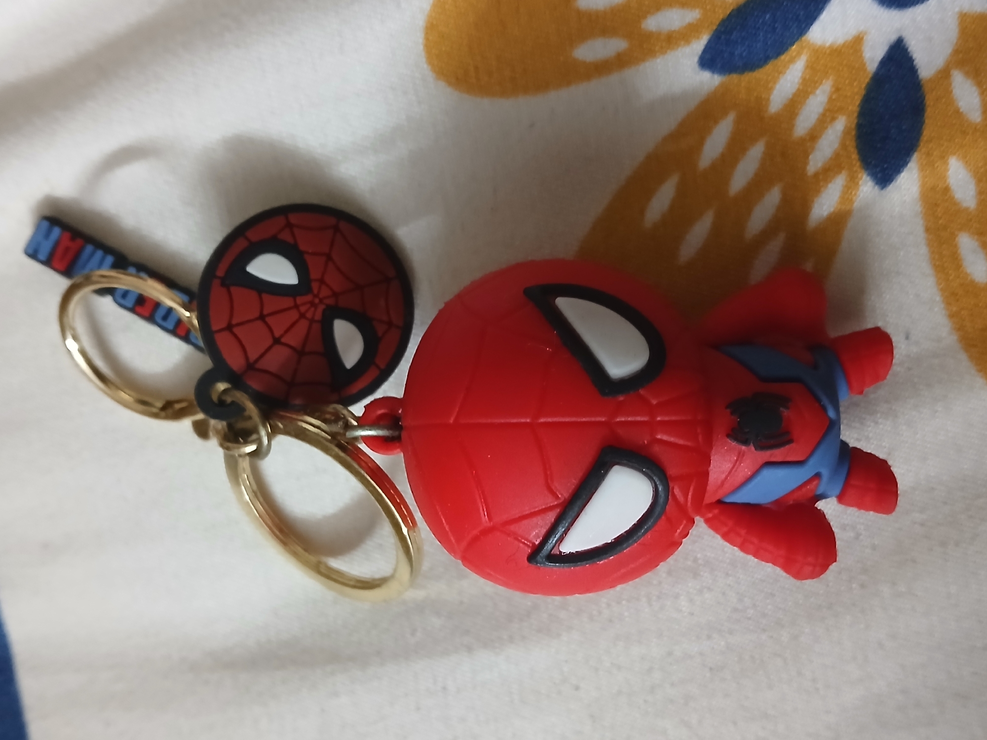 A close-up shot depicts a Spiderman-themed keychain lying on a patterned surface. The keychain features two main components: a round disc with a Spiderman mask design and a small Spiderman figure. Both are predominantly red with black and white detailing. They are connected by gold-colored metal rings and a small blue strap with the word "Spiderman". The background appears to be a patterned surface, likely a bedsheet or tablecloth, with white, yellow, and blue floral designs. The lighting is soft and diffused, creating subtle shadows. The shot is taken at an angle, providing a clear view of the keychain's details. The location is in the city of Bengaluru, India.