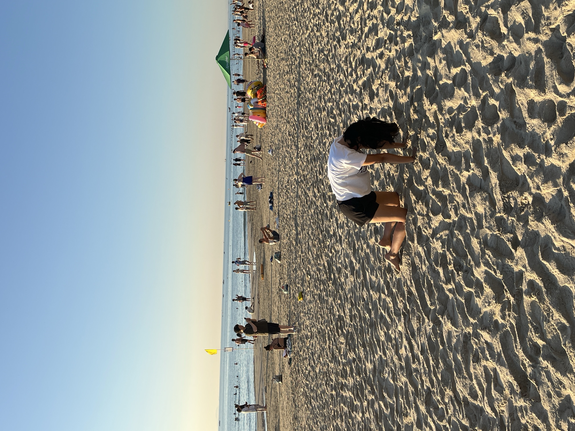 A beach scene in Đà Nẵng, Vietnam, shows numerous people enjoying a sunny day. The sky is clear, and the ocean is calm. People are swimming, sunbathing, and walking along the shoreline. The sand is light-colored and patterned with footprints and wave marks. In the foreground, a person kneels and leans forward, touching the sand with their hands. They wear a white shirt and black shorts. Further back, other beachgoers are visible, along with beach umbrellas, and inflatable toys. A yellow flag stands near the water's edge. A green tent is also visible further down the beach.