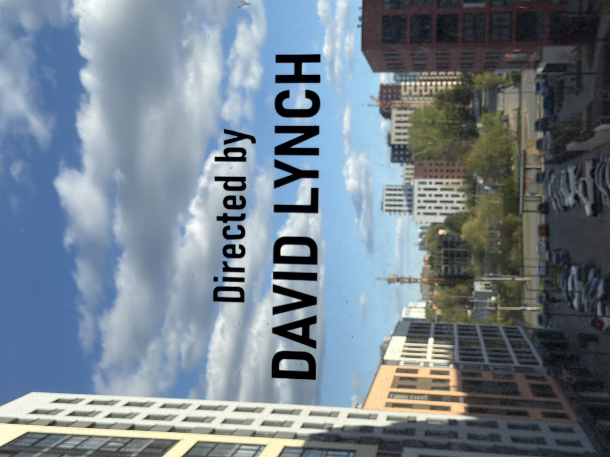 The image shows a view through a dirty glass window in the city of Izhevsk, Russia. The sky has a few clouds, and several buildings and a parking lot are visible. The text "Directed by DAVID LYNCH" is prominently displayed vertically over the scene.