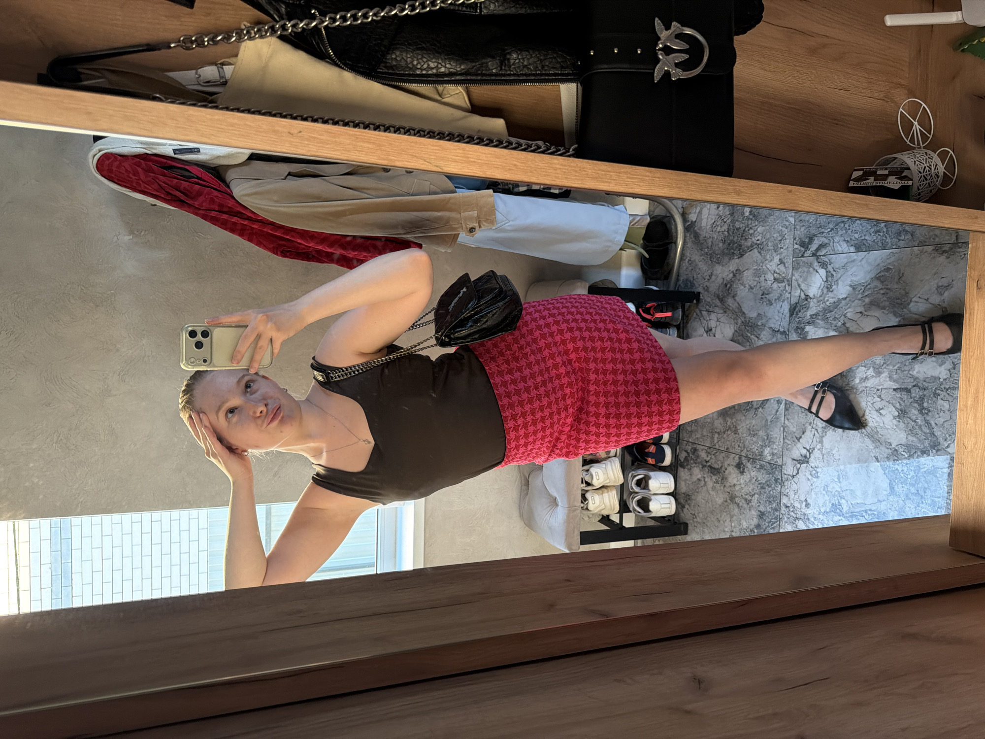 A young woman, approximately in her twenties, is captured in a mirror selfie within an indoor setting in Rossiyskiy, Russia. She holds a light-colored smartphone in her right hand, aiming it towards her reflection. Her left hand is placed on her head, and she has a slightly puckered lip expression while looking towards the phone. She wears a dark brown tank top, a short, fitted skirt with a pink and red houndstooth pattern, and black Mary Jane style shoes with two straps. A silver-toned chain necklace with a small pendant is visible around her neck, and a black shoulder bag with a chain strap is slung over her left shoulder.
The immediate surroundings visible in the reflection include a light gray textured wall, a white-framed window revealing an exterior brick wall, and a gray and white marble-patterned tiled floor. To the left of the reflection is a wooden door frame or panel. Behind the woman's legs, a small gray upholstered bench or ottoman sits above a black metal shoe rack holding several pairs of sneakers. To the right of the reflected woman, a garment rack or hooks display a red patterned jacket, a beige jacket, and a light blue shirt. A black leather-textured handbag with a distinctive "swallow" emblem, likely Pinko brand, hangs in the foreground on the right. Further to the right, on a wooden surface, a white Wi-Fi router, a small white wire-frame decorative bicycle ornament, and a white basket with papers are visible.