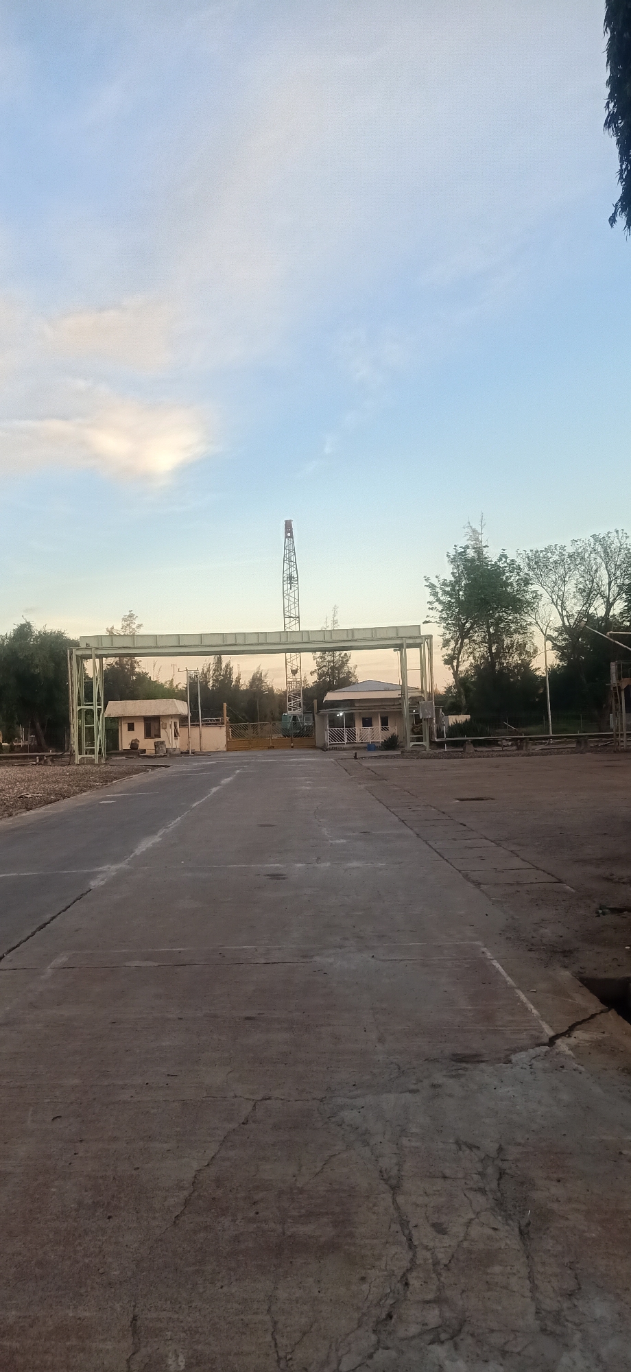 The image depicts an outdoor scene with a wide, paved road leading towards a green archway. To the left and right of the road, under the archway, are small, light-colored buildings. Beyond the archway, a yellow gate is visible, and in the distance, a tall construction crane stands against a pale blue sky with scattered clouds. Trees and other vegetation surround the area. The overall impression is that of an industrial or construction site entrance. The scene does not contain any people, actions, or interactions. The location is Palohlada, Indonesia.