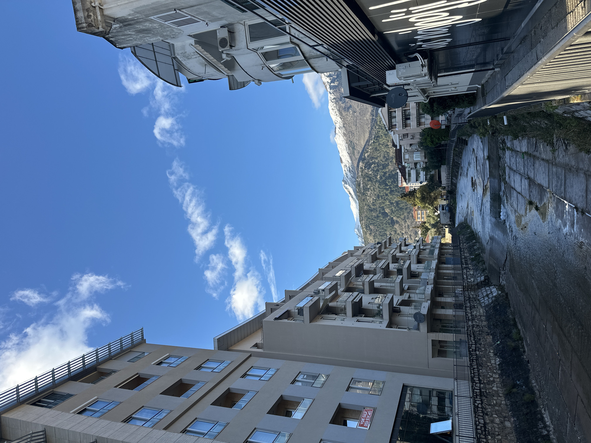 The image depicts a view in Budva, Montenegro. The frame is divided by buildings and a mountain range in the background. On the left, there are modern buildings of multi-story residential complexes. The buildings are characterized by rectangular windows and protruding balconies. A "FOR SALE" sign is visible in one window. Above the buildings is a bright blue sky with scattered white clouds.
On the right, there is a partially visible, snow-covered mountain range. In the foreground, there is a canal with water flowing through it, and the edge is defined by concrete stones and vegetation. The buildings above the canal are a mix of residential and commercial structures. The light is bright, suggesting a clear day.