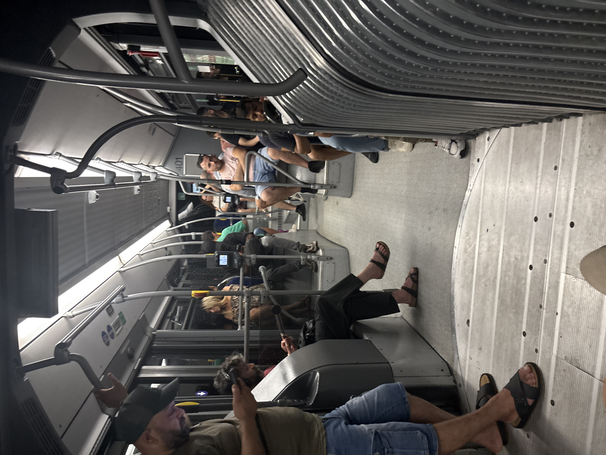 The image shows the interior of a public bus, likely in Venice, Italy, given the context. The bus is moderately crowded with passengers seated and standing. The lighting is artificial, with overhead lights illuminating the aisle and seats. The aisle flooring is a light grey, with visible metal fasteners. Along the right side of the bus, a corrugated metal barrier is prominent.
In the foreground, on the left, a man wearing a dark baseball cap, a khaki-colored t-shirt, and denim shorts is looking down at his phone, holding it in his left hand. His right hand is on a handrail. Beside him, a person in dark pants and sandals is seated, looking at their phone.
Further down the aisle, several other passengers are visible. A woman with blonde hair wearing a patterned dress is seated on the left. In the middle of the bus, a man in a pink t-shirt and denim shorts is seated with his arms crossed. Behind him, several other passengers are seated and standing, some looking at their phones. The general atmosphere suggests a typical commute. The bus number "401" is visible on a sign in the background.