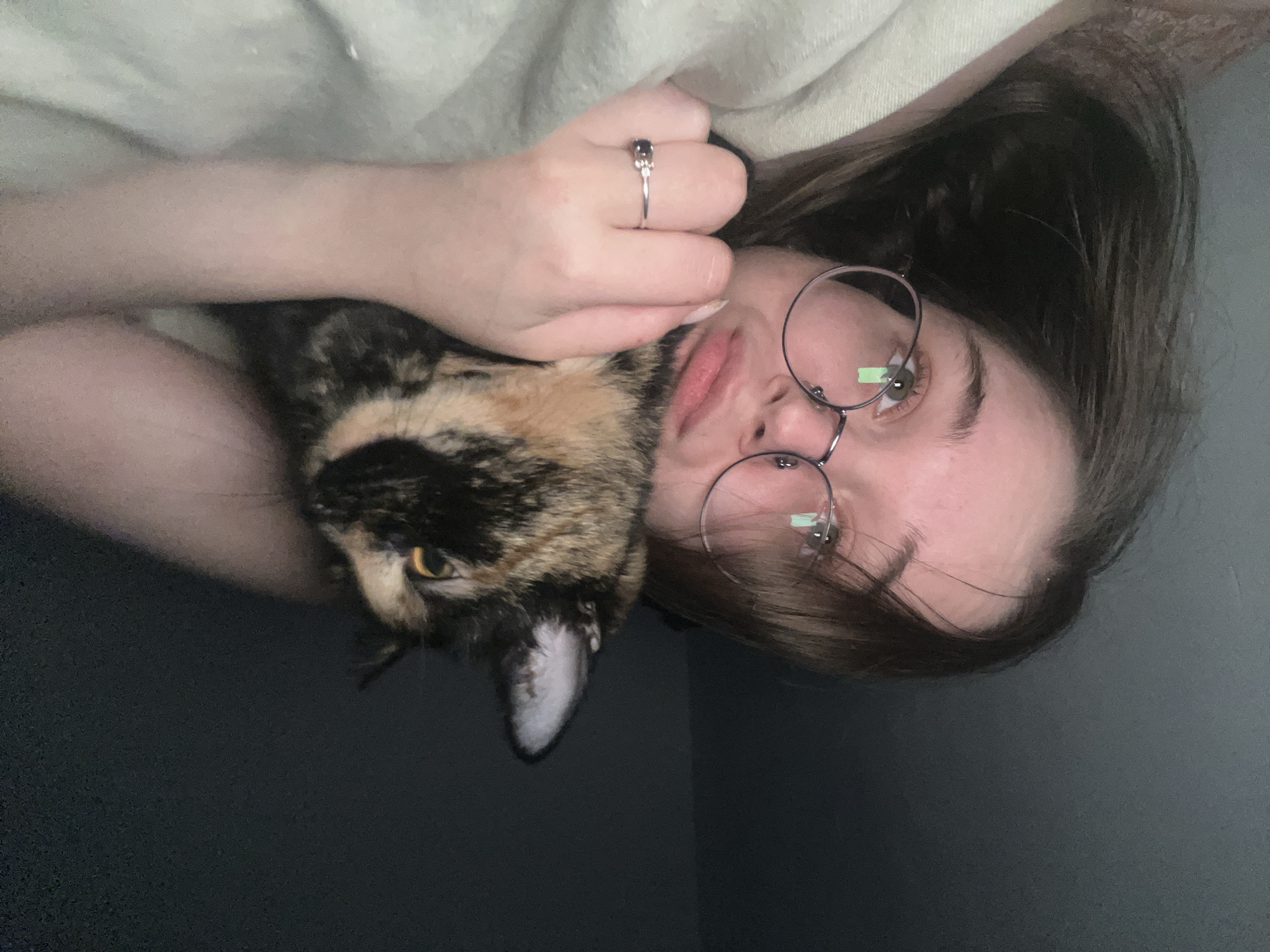 A woman with long brown hair and glasses is lying down, cradling a calico cat. The woman is wearing a silver ring on her left hand. The background is a mix of dark and light, likely indicating a room with some lighting. There is no information suggesting this image was taken in Pushchino, Russia.