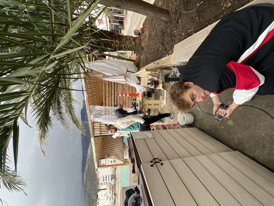 The image captures an outdoor construction or renovation scene by the sea in Budva, Montenegro, under an overcast sky with dark clouds visible over distant mountains.
Three adult males are present. In the foreground, a male with light brown, curly hair, wearing a black, red, and white jacket, squats on a concrete surface. He holds a black smartphone horizontally in both hands, angled towards the right, and looks directly at the camera. A black cable extends from the smartphone or its vicinity.
In the midground left, a male with a man bun and beard, dressed in a light-colored jacket over a green t-shirt and dark pants, stands with his left hand in his pocket, looking towards the left side of the frame.
In the midground center-right, a third male, wearing a dark shirt and dark pants, is bent over a wooden workbench, actively engaged with power tools. A red and white striped warning cone stands adjacent to the workbench.
The immediate foreground features a light-colored, horizontally slatted composite wood counter or partition with black cabinet handles. On its surface, a black and red circular saw and a white ceramic cup are visible. A white bucket and a metal beer keg are positioned on the ground near the counter.
Behind the individuals, a semi-permanent wooden structure with a woven canopy roof and transparent plastic sheeting, indicative of ongoing work, is present. This structure faces a body of turquoise-green water. Multi-story buildings line the distant coastline. A large palm tree with prominent fronds occupies the right side of the frame, with exposed soil at its base.