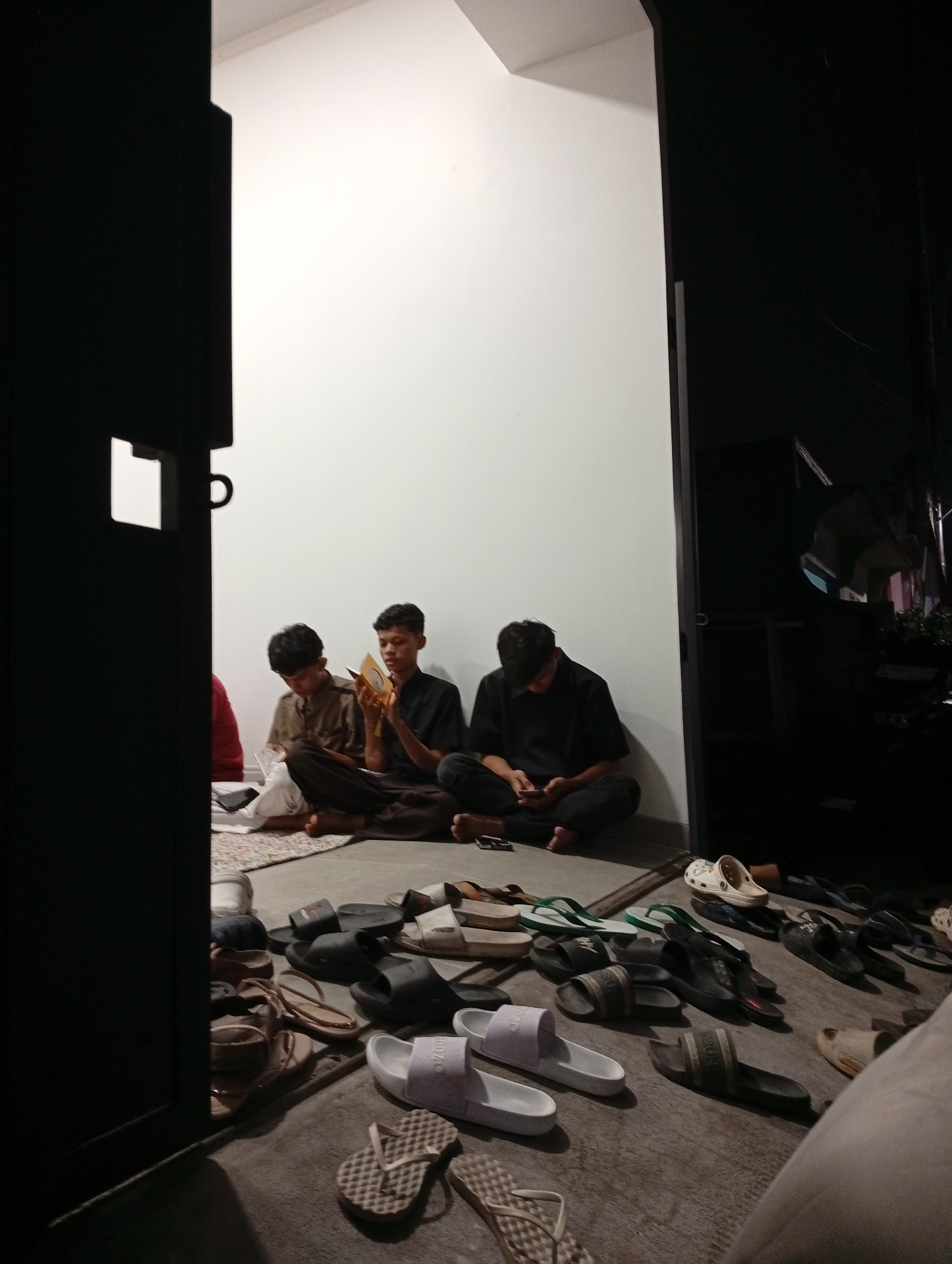 Three young men are seated on the floor in front of a white wall, possibly in the city of Caringin, Indonesia. Two of the men are looking at a book or religious text. The third man is looking at his phone. There are several pairs of sandals on the floor in front of them. The scene appears to be indoors, with a dark doorway visible on the right side of the image.