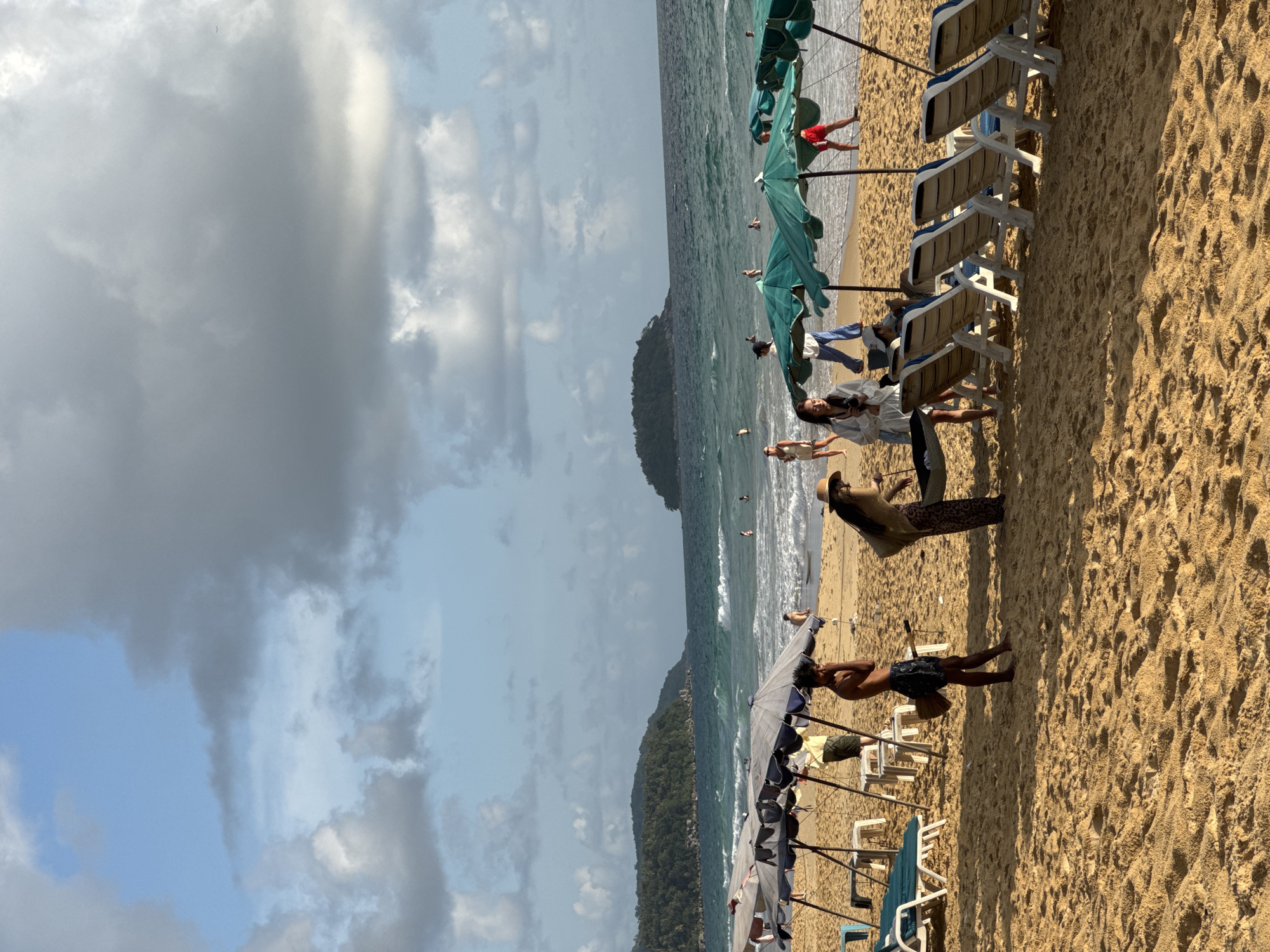 In Karon, Thailand, a wide sandy beach is depicted under a partly cloudy sky. The ocean exhibits moderate waves and a turquoise hue, with several people swimming or wading in the water. Along the shoreline, numerous beach chairs are set up, some occupied by individuals, and umbrellas provide shade.
In the foreground, a man in patterned shorts stands near a beach umbrella, holding a broom. To his right, a woman in a wide-brimmed hat and patterned skirt leans over. Further back, a group of people stand and converse near more umbrellas and chairs. In the middle ground, several individuals are enjoying the water. The background features a distant island covered in lush green vegetation, with the sea stretching to the horizon. The overall scene captures a relaxed atmosphere on a popular tourist beach.