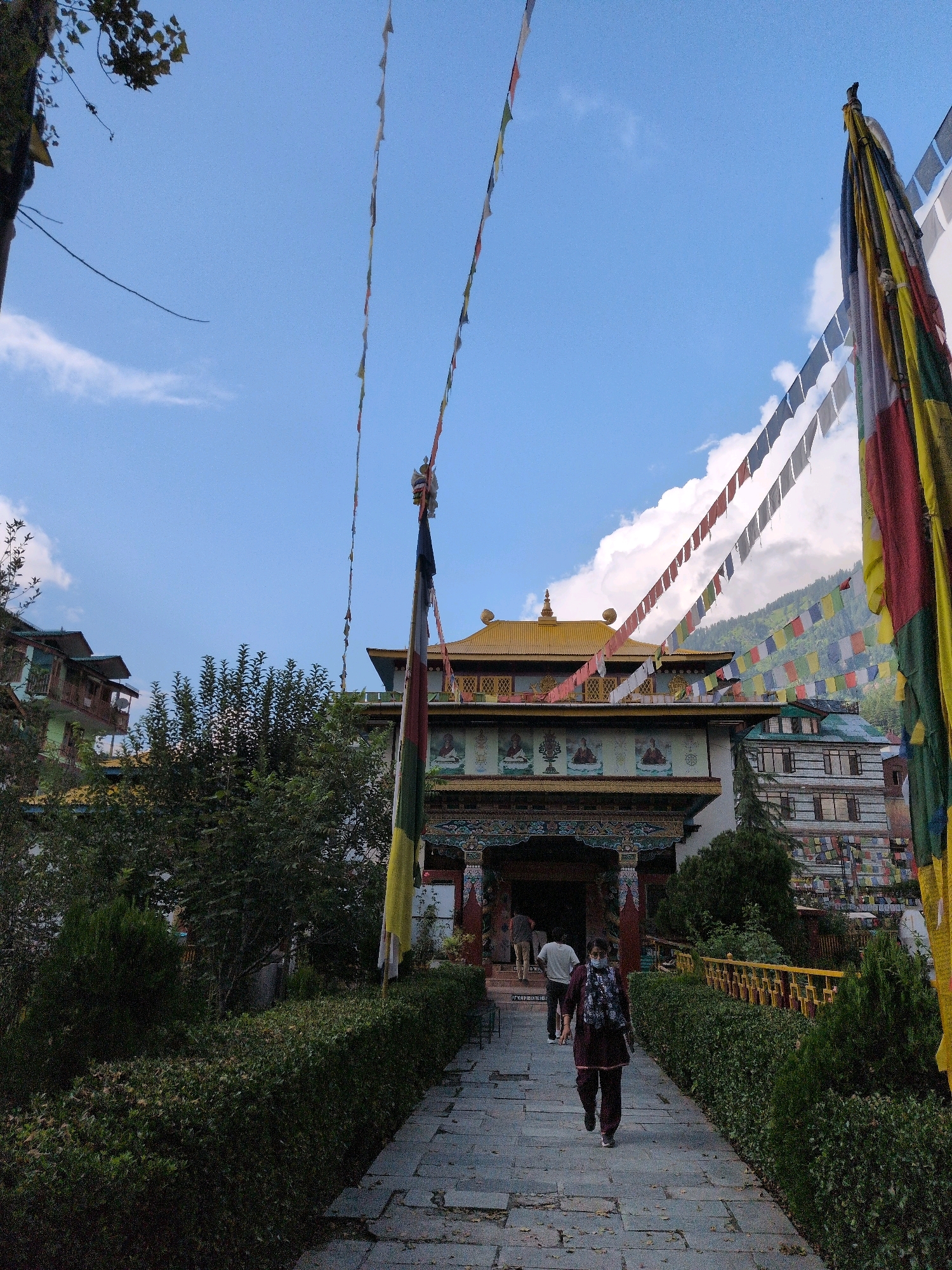 The image depicts the entrance to the Tibetan Monastery in Manali, India. The monastery features a traditional architectural style with a yellow, multi-tiered roof and intricate carvings. Prayer flags, in various colors, are strung across the sky, adding to the spiritual ambiance. A paved walkway leads to the monastery entrance, flanked by manicured hedges and trees. Several individuals are visible, including two people walking towards the entrance and one person walking away from it. The sky is partly cloudy, with a range of green hills in the background, suggesting the monastery is situated in a mountainous region. The scene captures a serene moment at a significant cultural landmark in Manali.