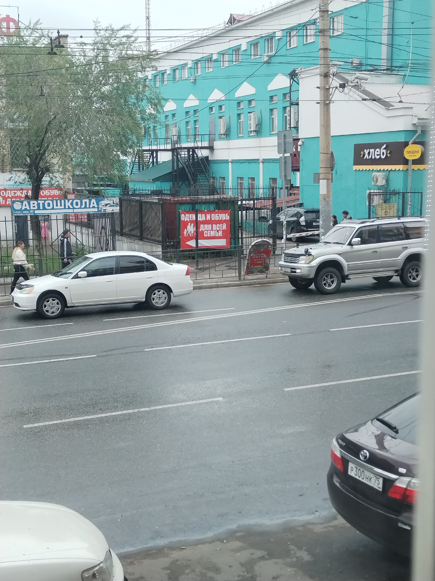 A street scene in Chita, Russia, features a turquoise and white multi-story building with various signs. On the left, a white sedan is parked near a fence. To the right of the sedan, a silver SUV is on the road. In the foreground, the rear of a dark sedan is visible, displaying a license plate with the characters "P300HK75". The road appears wet, indicating recent rain or damp conditions.