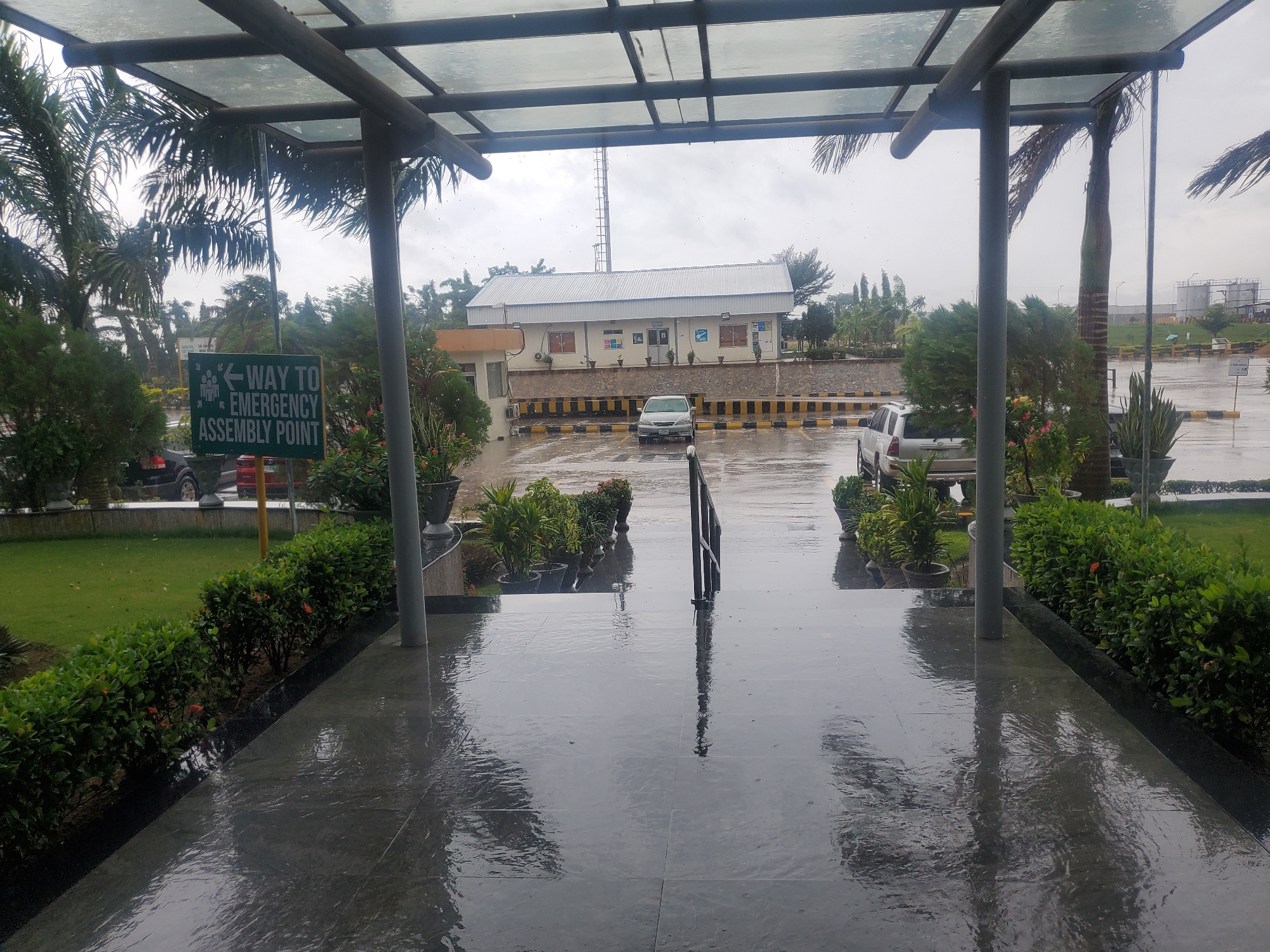 This image depicts a rainy day scene from under a covered walkway at what appears to be an industrial or corporate facility in Gwagwada, Nigeria. The walkway has a transparent roof supported by dark metal beams. The wet, dark tiled floor of the walkway reflects the overhead structure and the overcast sky.
To the left, a green sign with white text reads "WAY TO EMERGENCY ASSEMBLY POINT" with a directional arrow and an icon of people. Beyond the sign, a dark-colored vehicle is partially visible. Lush green grass and manicured bushes with small orange flowers line the walkway's edge. Palm trees with fronds blowing in the wind are visible behind the bushes.
In the middle ground, a light-colored sedan is parked in a lot with yellow and black striped barriers. Behind the parking lot, a white building with a metal roof is visible, along with a tall communications tower in the distance. Other vehicles, including a silver SUV, are present in the lot. The ground in the parking area is also wet and appears to have standing water.
To the right, more potted plants and green bushes are seen, along with a dark grey vertical pole. In the background, industrial structures and more greenery are visible under a grey, cloudy sky. The overall impression is of a functional outdoor space in a tropical climate experiencing inclement weather.