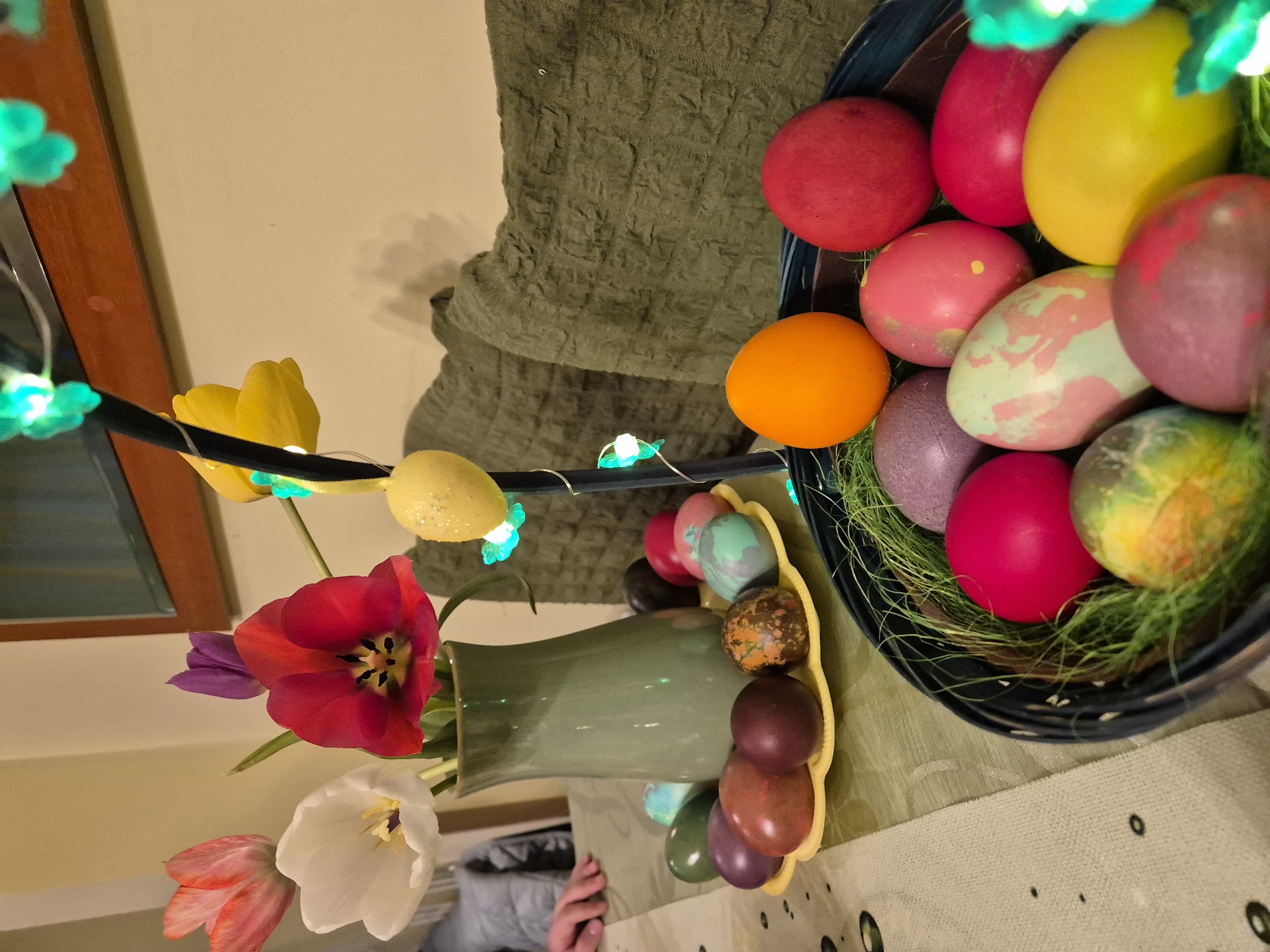 A close-up shot taken indoors features two arrangements centered around Easter. The arrangement on the right consists of a blue basket filled with dyed Easter eggs of various colors (red, yellow, pink, orange, purple, green) nestled in green faux grass. The other arrangement, on the left, includes a green vase containing tulips (red, white, pink, purple) and yellow Easter eggs. A string of green clover-shaped lights is draped over these arrangements. The background is a beige wall and a textured green cloth. A partial view of a person's hand can be seen near the base of the vase. The table is covered with a light-colored cloth with a subtle pattern. The location is Unknown, Unknown.