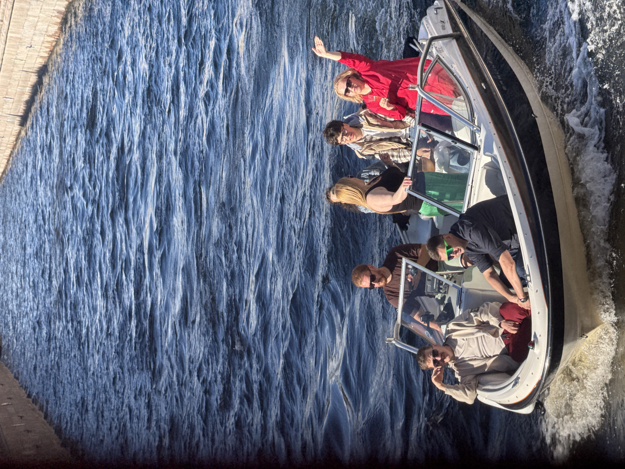 A group of eight people are enjoying a boat ride on a sunny day. The boat, a white motorboat with a dark stripe, is moving through choppy blue water, creating white spray. The water appears to be a river or canal, given the presence of a stone embankment on the left side of the image. The people are of various ages and genders. Two individuals are at the front of the boat, one in a red top waving, and another in sunglasses. Several other people are seated and standing further back in the boat, some appearing to be talking and laughing. The overall mood seems relaxed and joyful, with people enjoying the sunshine and the ride. There is no visible text in the image.