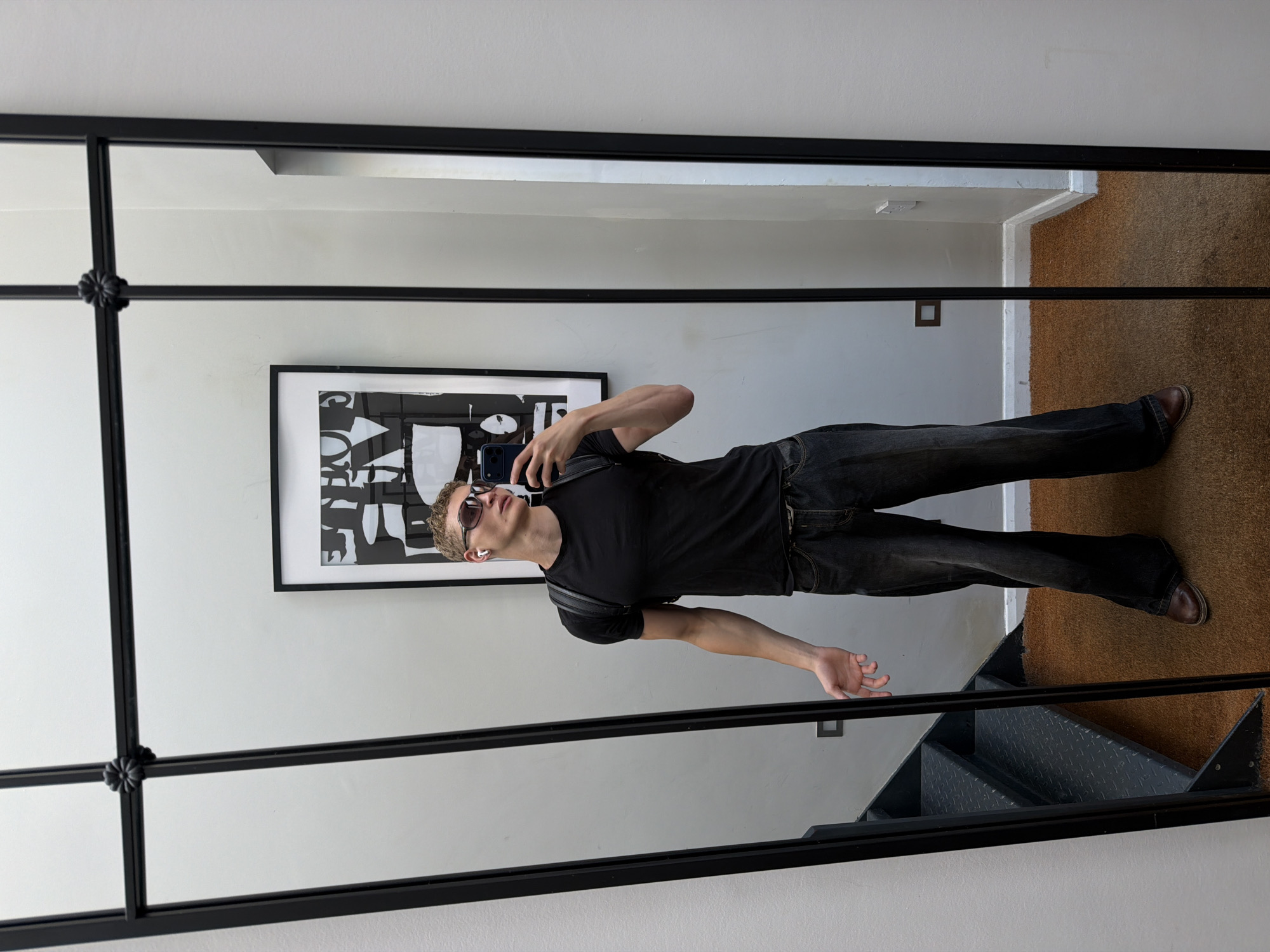 A man is taking a mirror selfie in a room. He is wearing a black t-shirt, dark wash jeans, and brown shoes. He has on sunglasses and white earbuds. He is holding a black smartphone to take the picture. In the background, there is a framed piece of abstract art on the wall. The room has a light-colored wall and a brown carpet. A metal staircase is visible to the right of the man, suggesting a multi-level space. The mirror itself is framed with a black metal border, divided into several panes by horizontal and vertical mullions. The lighting suggests it is daytime, and there are no clear indications of the weather outside. The man's pose and expression are casual and relaxed. There is no visible text in the image. The location is London, United Kingdom.