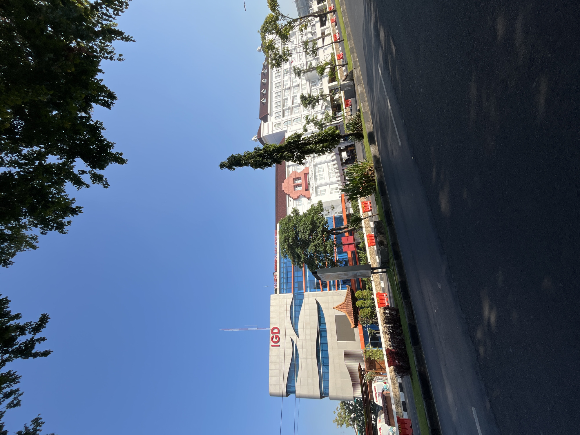 This is a daylight, eye-level, medium shot of a hospital complex on a clear blue sky day in Sidoarjo, Indonesia. The foreground features a black asphalt road with a white line marking, reflecting dappled sunlight. The middle ground displays a row of green grass and plants bordering the road, with red and white safety barriers. Behind the barriers, a modern hospital building with a gray, curved facade and blue-tinted windows is visible, displaying "IGD" in red letters. Adjacent to it is a white, multi-story building with arched windows and a red-tiled roof. Several trees, including a tall, slender evergreen and a leafy deciduous tree, are scattered throughout the hospital grounds. A small, ornate gazebo-like structure with a red-tiled roof is situated in front of the modern hospital section. An ambulance is partially visible on the left. In the background, the sky is clear and blue with the tops of trees framing the scene from the left and right edges.