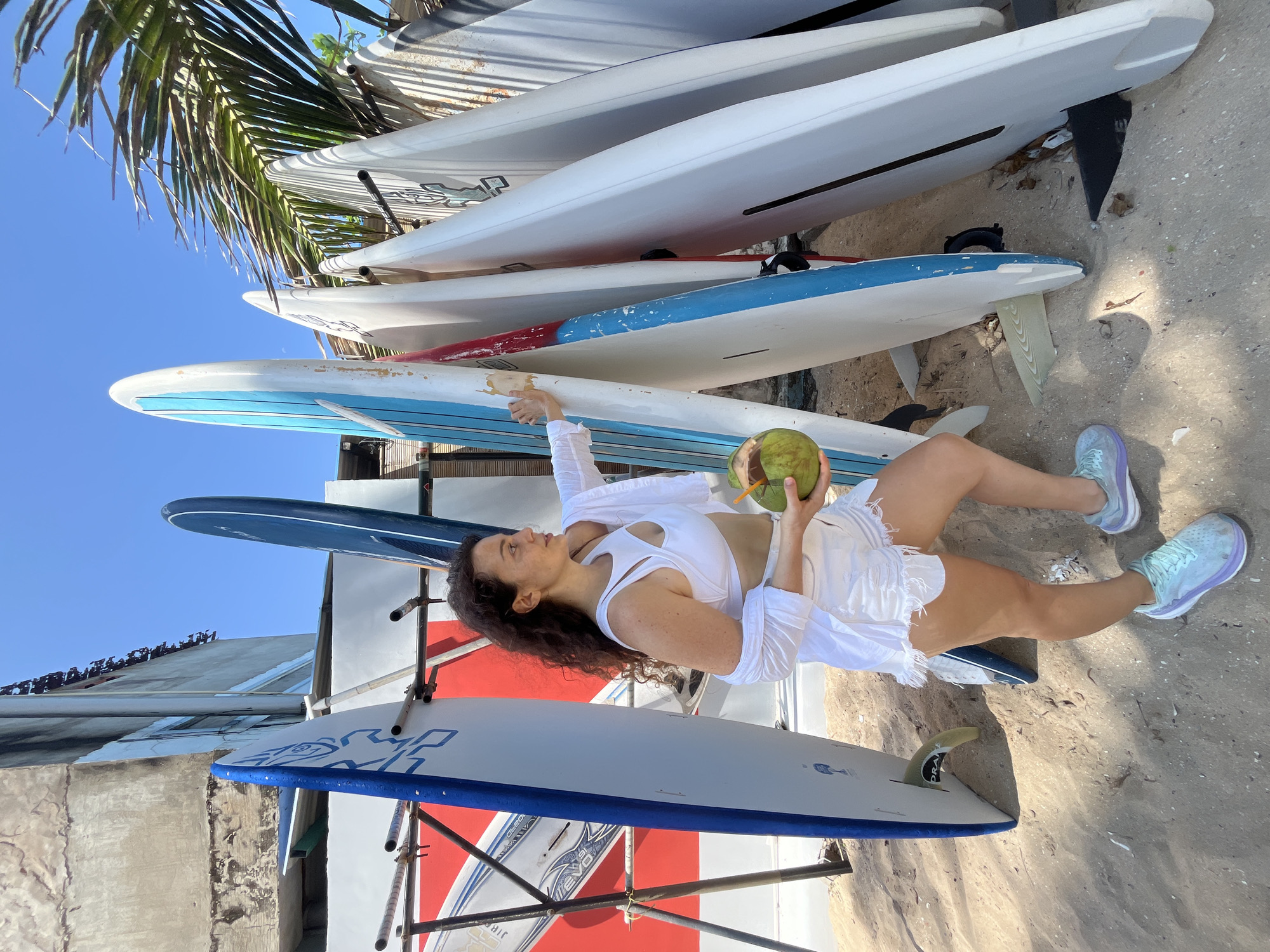 A woman in white athletic attire holds a halved coconut with a straw in it while standing on a sandy beach in Phan Thiet, Vietnam. She is surrounded by a rack of surfboards, with some leaning against a building. The woman is positioned between two surfboards, one on her left and another on her right. The background shows a building with weathered concrete walls and a clear blue sky with palm fronds from a tree visible. The woman is looking to her right, her left hand extended towards one of the surfboards. The scene is outdoors and appears to be during daylight.
