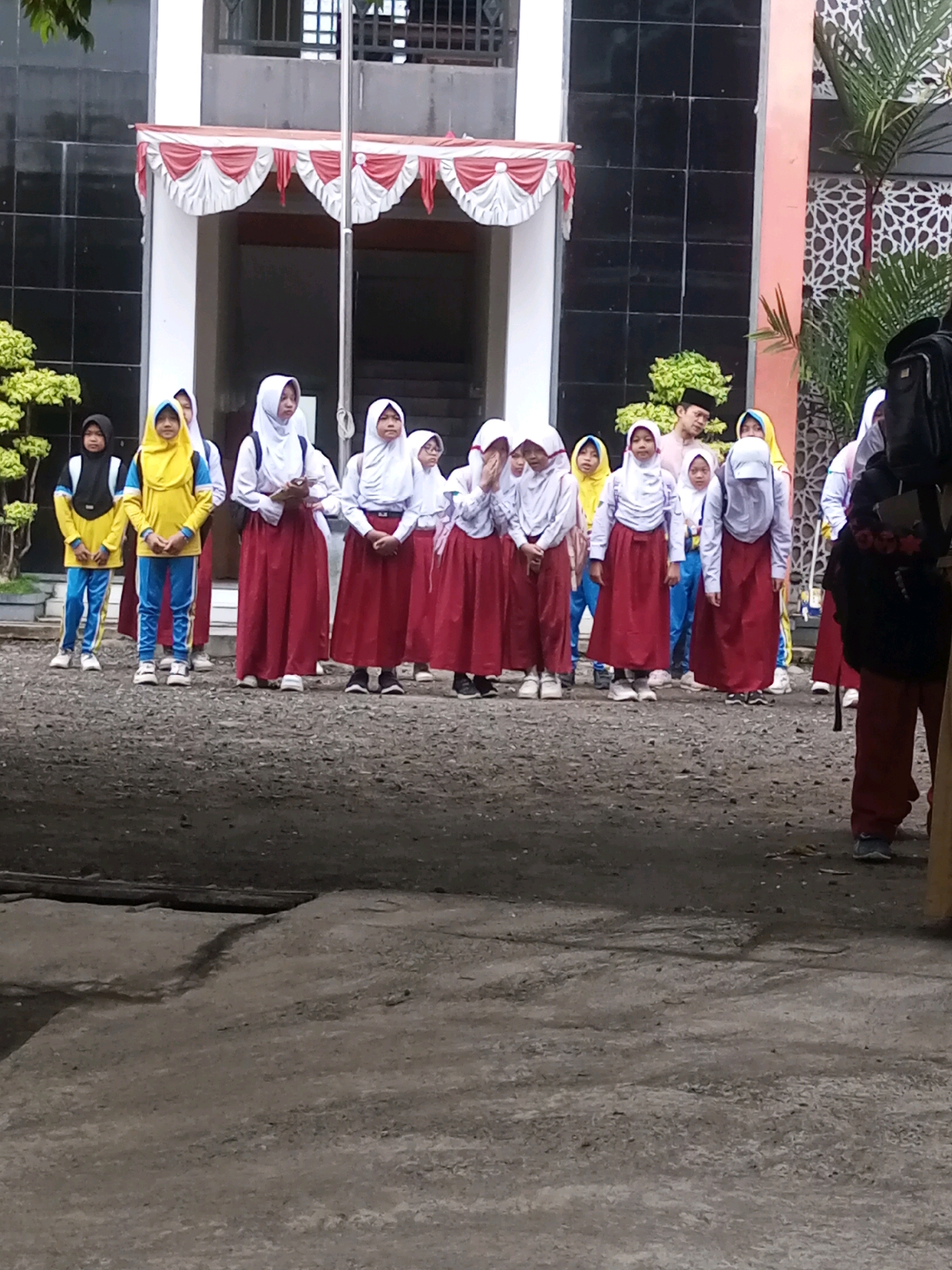 A group of students in uniform are standing on a paved area in front of a building. The students are mostly wearing white hijabs and red skirts, with some wearing yellow shirts and blue pants. A man in a pink shirt and black peci is standing behind them. The building in the background has a tiled facade and a white and red banner above an entrance. The ground is covered in gravel and dirt. The image appears to have been taken in an outdoor educational setting.