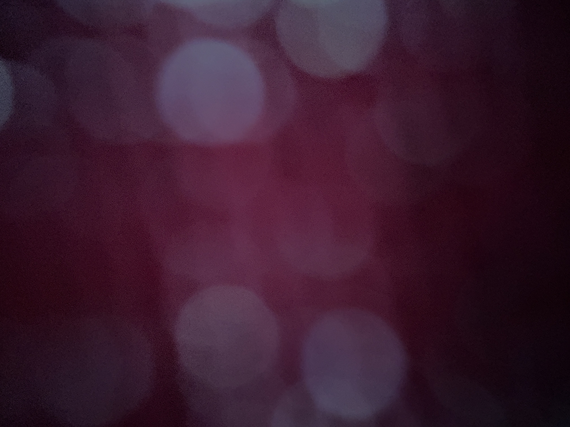 This is an abstract image characterized by an out-of-focus, bokeh effect. The dominant colors are deep maroon, dark red, and hints of purple in the background, which appears textured due to the blurring.
**Subjects:** There are no discernible people or distinct objects in the image. The main subjects are abstract circular shapes of light, ranging from bright white to soft grey and faint purple, created by the bokeh effect. These light sources vary in size and intensity, scattered across the frame, with larger, brighter circles prominently featured in the upper and lower halves.
**Activity or event taking place:** No specific activity or event is depicted due to the abstract and blurred nature of the image. It represents an artistic or atmospheric portrayal of light and color.
**Notable details:**
- **Time of day:** The dark background and prominent bokeh lights suggest an evening or nighttime setting, or an indoor environment with low ambient light.
- **Weather:** Not discernible.
- **Location cues:** There are no specific location cues within the image itself to indicate Corfu, Greece, or any other place. The blurred background could be a wall, fabric, or distant lights.
- **Emotions:** The soft focus and warm, dark tones could evoke feelings of calm, contemplation, or a cozy, ambient atmosphere.
- **Visible text:** No visible text is present.