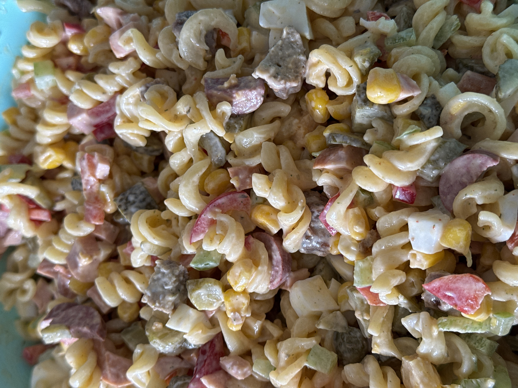 The image shows a close-up, overhead view of a bowl filled with pasta salad. The pasta is fusilli-shaped and coated in a creamy dressing. Mixed in with the pasta are various ingredients, including diced red bell pepper, green pickles, corn kernels, and chunks of what appear to be hard-boiled egg and processed meat, possibly ham or sausage. The salad is densely packed in a light blue bowl, only the rim of which is visible at the top. There is no indication of the location in Siegburg, Germany, or any environmental context beyond the food itself.