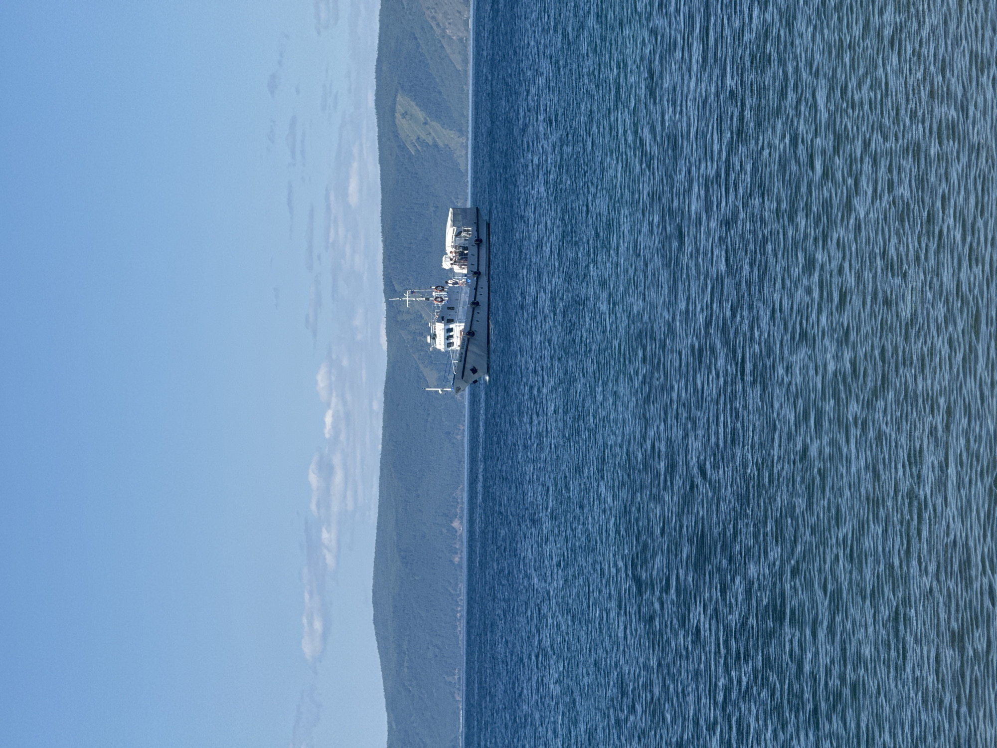 A white boat with a red stripe sails on the blue water of Lake Baikal, near Listvyanka, Russia. The boat has several people on its deck. In the background, a range of green, tree-covered hills rises under a clear blue sky with a few wispy clouds. The water has small ripples across its surface.