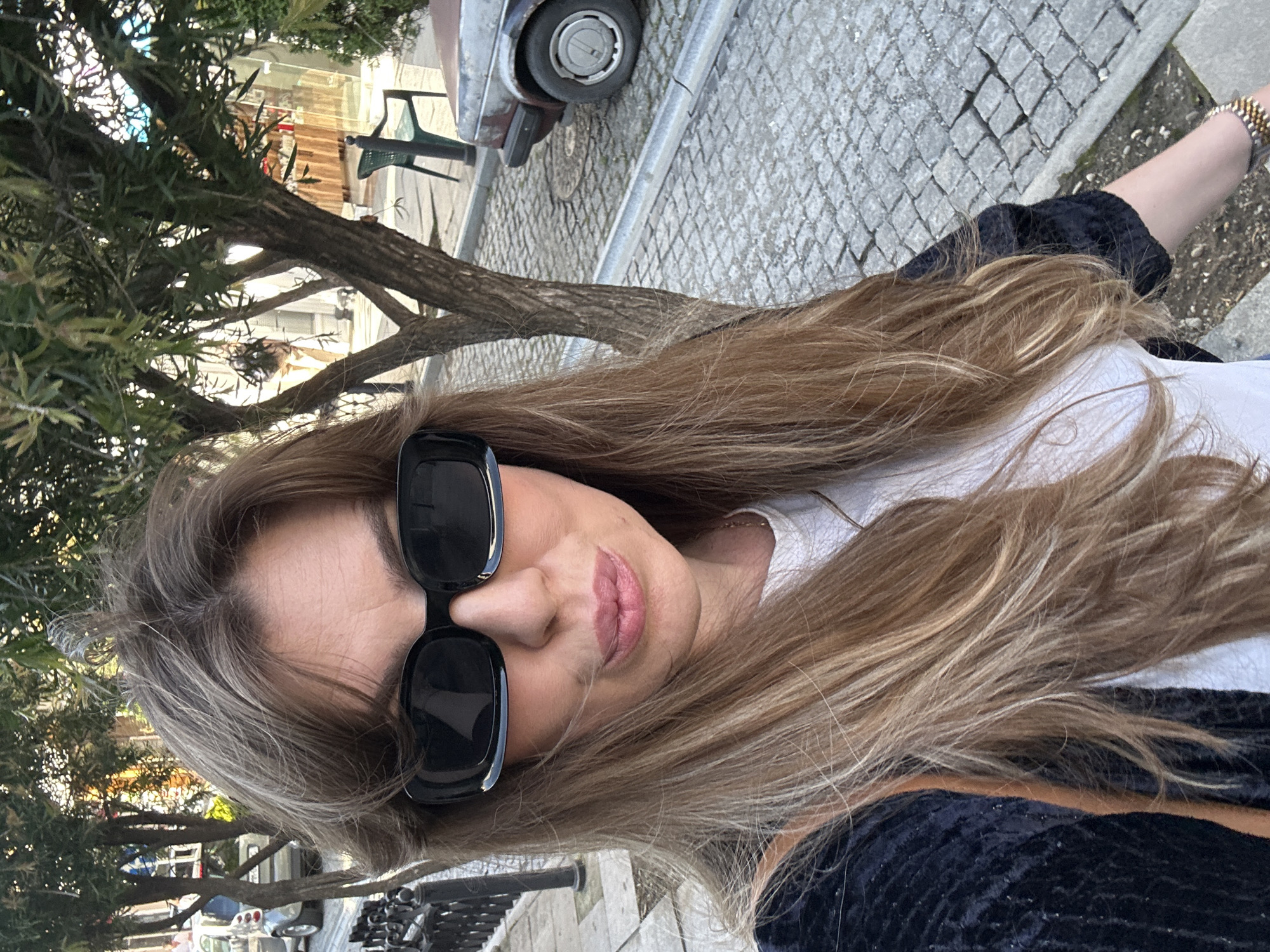 A woman with long, light-brown hair is taking a selfie outdoors. She is wearing large, black sunglasses and a white t-shirt under a black jacket. She is standing on a cobblestone street next to a sidewalk with a metal railing. Behind her, there is a tree and a partially visible old car. The location is in Batumi, Georgia.