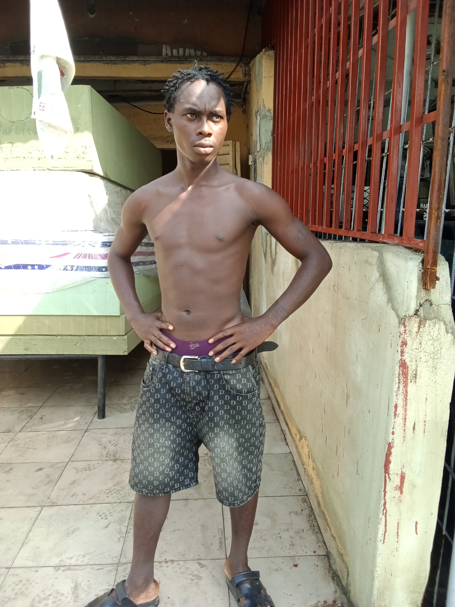 A young, dark-skinned man stands shirtless in an outdoor setting, likely a courtyard or passage in Ado, Nigeria, under bright daylight. He faces slightly right, looking off-camera with a neutral expression. His short, dark hair is styled in small twists or dreadlocks. He wears dark shorts patterned with repeating white "D" letters, a black belt with a silver buckle, and visible purple underwear. His hands are placed on his hips, and he wears dark sandals or slides.
To the left of the man, several mattresses, some wrapped in plastic and featuring red and blue patterns, are stacked on a light green base. A black metal leg from a bed frame is visible beneath them. Behind the man and extending to the right is a light-colored concrete wall, showing signs of wear, chipping paint, and some reddish stains or streaks. Further to the right, a red metal gate or railing with vertical bars is visible. The ground is covered with light-colored tiles, marked with various stains and water spots. Bright sunlight illuminates the scene, casting distinct shadows. There's blurry text on the mattress wrapping ("EE" and patterns) and on a structure behind the gate ("HAP" likely part of "HAPPY").