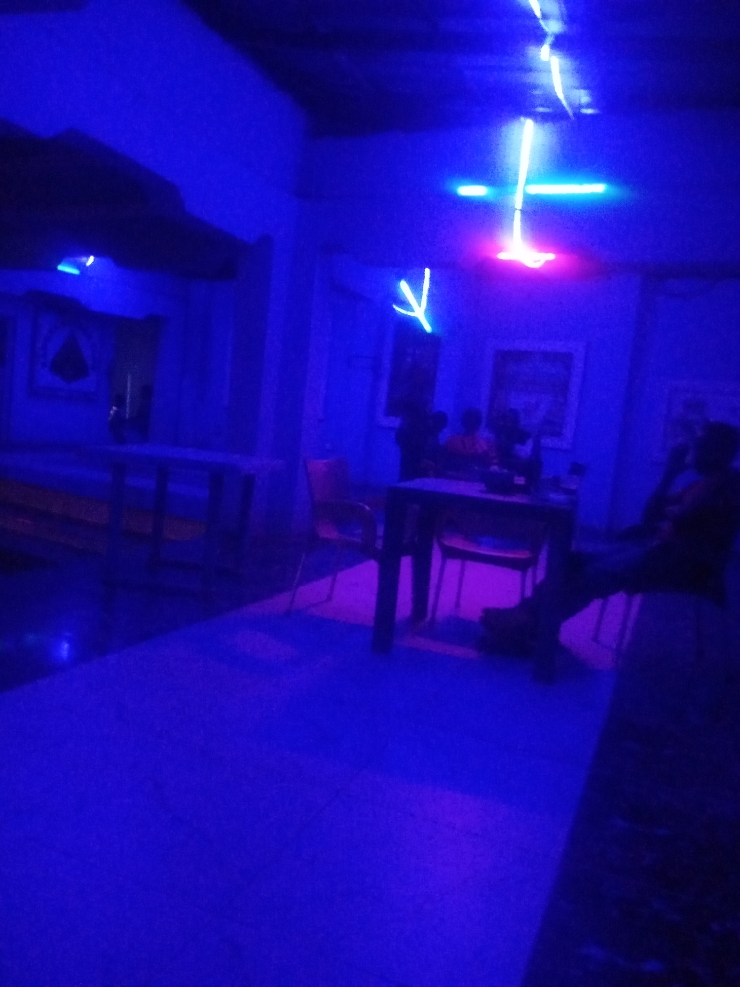 The image depicts a dimly lit indoor space, likely a bar or lounge, illuminated by vibrant blue and pink neon lights. In the foreground, a section of the floor is visible, reflecting the colorful lighting. Several tables and chairs are present, suggesting a seating area. Some individuals are visible in the background, appearing as silhouettes due to the low light conditions. One person is seated at a table, and others are standing or interacting. The overall ambiance suggests a lively or social gathering. The presence of paintings on the wall and the arrangement of furniture indicate a deliberate establishment for public use. The scene is consistent with an entertainment venue located in Sabon Gari, Nassarawa, Nigeria, known for its vibrant nightlife.