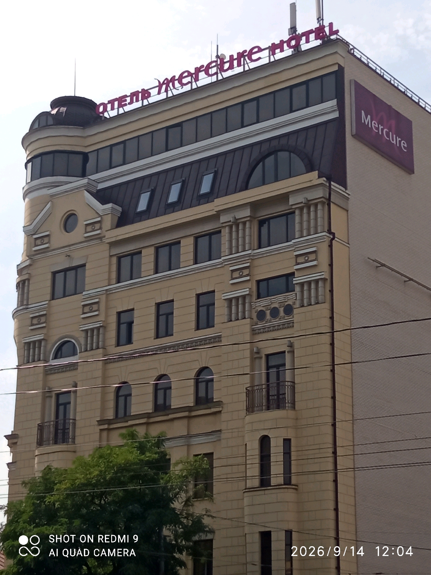 The image shows a close-up view of the Mercure hotel building in Rostov-on-Don, Russia. The hotel has a light yellow facade with ornate architectural details, including columns and decorative elements around the windows. The upper part of the building features a dark-colored roof with skylights, and a prominent sign with the hotel's name "Mercure" in purple lettering is visible on the side of the building. The sky is overcast, and there are some power lines in the foreground. The bottom of the image displays a watermark indicating "SHOT ON REDMI 9 AI QUAD CAMERA" and the date "2026/9/14 12:04".