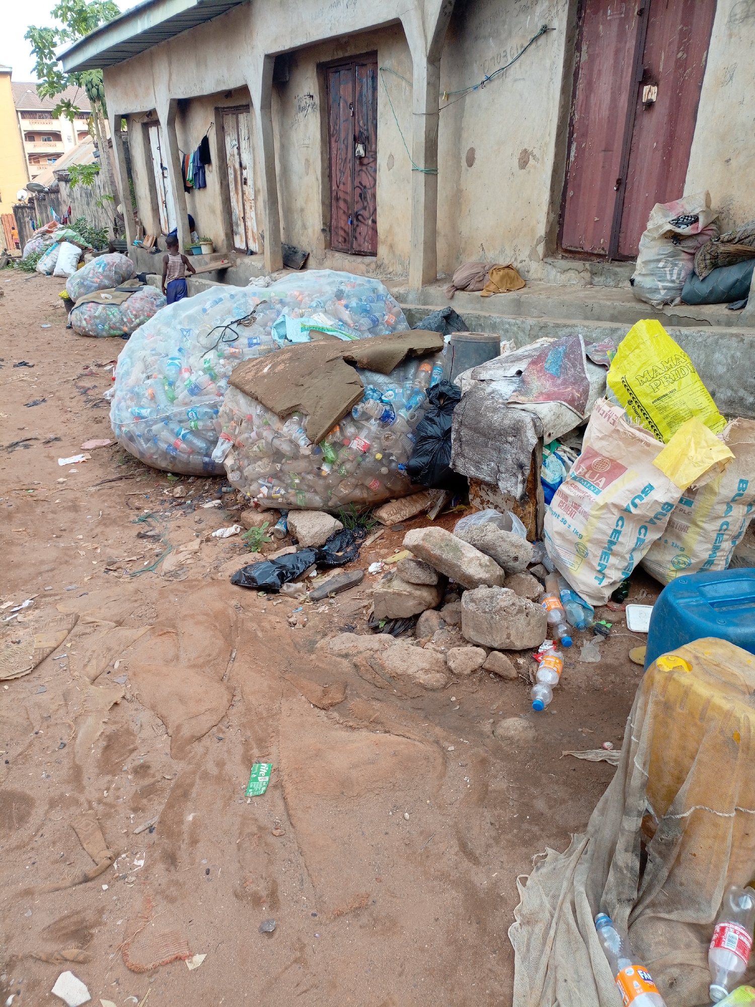 The image depicts an outdoor scene, likely an unpaved area or alleyway, situated alongside a weathered, single-story building. The ground is composed of reddish-brown dirt and sand, scattered with debris including empty plastic bottles, small rocks, and other waste.
The main subjects are several large, clear plastic bags, overfilled with numerous empty plastic bottles, indicating a collection point for recycling. These immense bags are prominent in the foreground and mid-ground. A young child, dressed in a striped shirt and blue shorts, stands near one of the building's doorways in the mid-ground, while another child's head is faintly visible further back.
The building's facade is unpainted concrete or stucco, displaying signs of wear and age, with multiple entrances framed by archways leading to rusty, dark red doors. Clothes are visible hanging near one of the doorways. To the right in the foreground, a heap of items includes more empty plastic bottles, concrete blocks, a blue and a yellow plastic jerrycan, and several white and yellow sacks. Text visible on these sacks includes "BUA CEMENT PLC", "PORTLAND-LIMESTONE CEMENT Net wt 50 ± 1kg", and "MAMA PRIDE". A green wrapper bearing "rite" is discernible on the ground.
The scene takes place during daylight hours, under natural light. It portrays a low-income or informal settlement setting, likely in a developing country, where plastic waste collection is a significant activity.