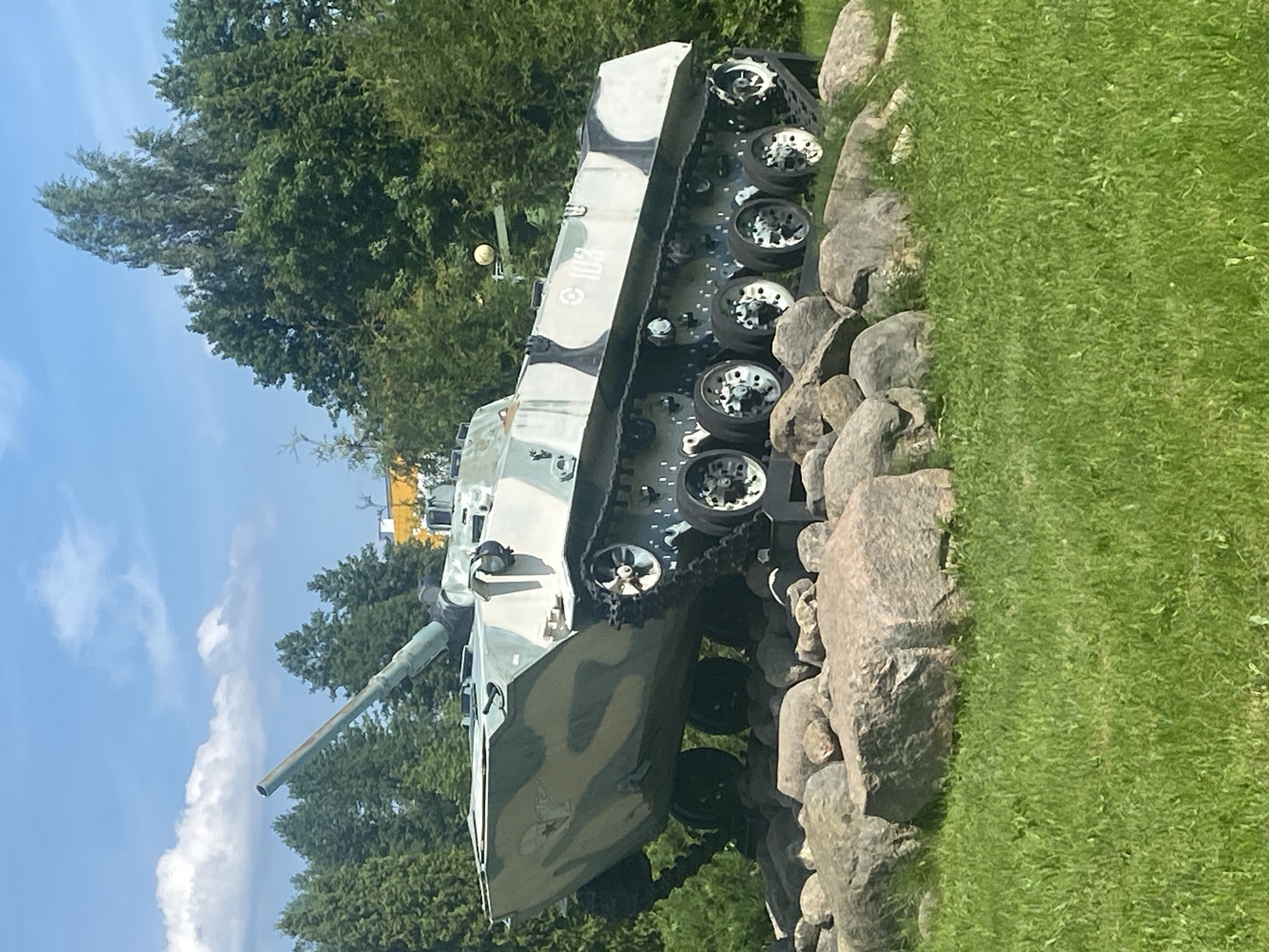 The image shows a parked, camouflaged military tank, likely a memorial or display, situated on a pile of large, gray rocks. The tank is positioned outdoors on a grassy area, with a backdrop of dense green trees under a blue sky with scattered white clouds. A yellow building is partially visible behind the trees on the right side of the frame. The tank is oriented at an angle, with its barrel pointing upwards and to the left. The camouflage pattern consists of irregular shapes in shades of green, white, and gray. The side of the tank features a white marking of "103" inside a white square border. The foreground is dominated by bright green grass. The scene appears to be a public space in Krupki, Belarus, given the context provided. There are no people visible in the image.