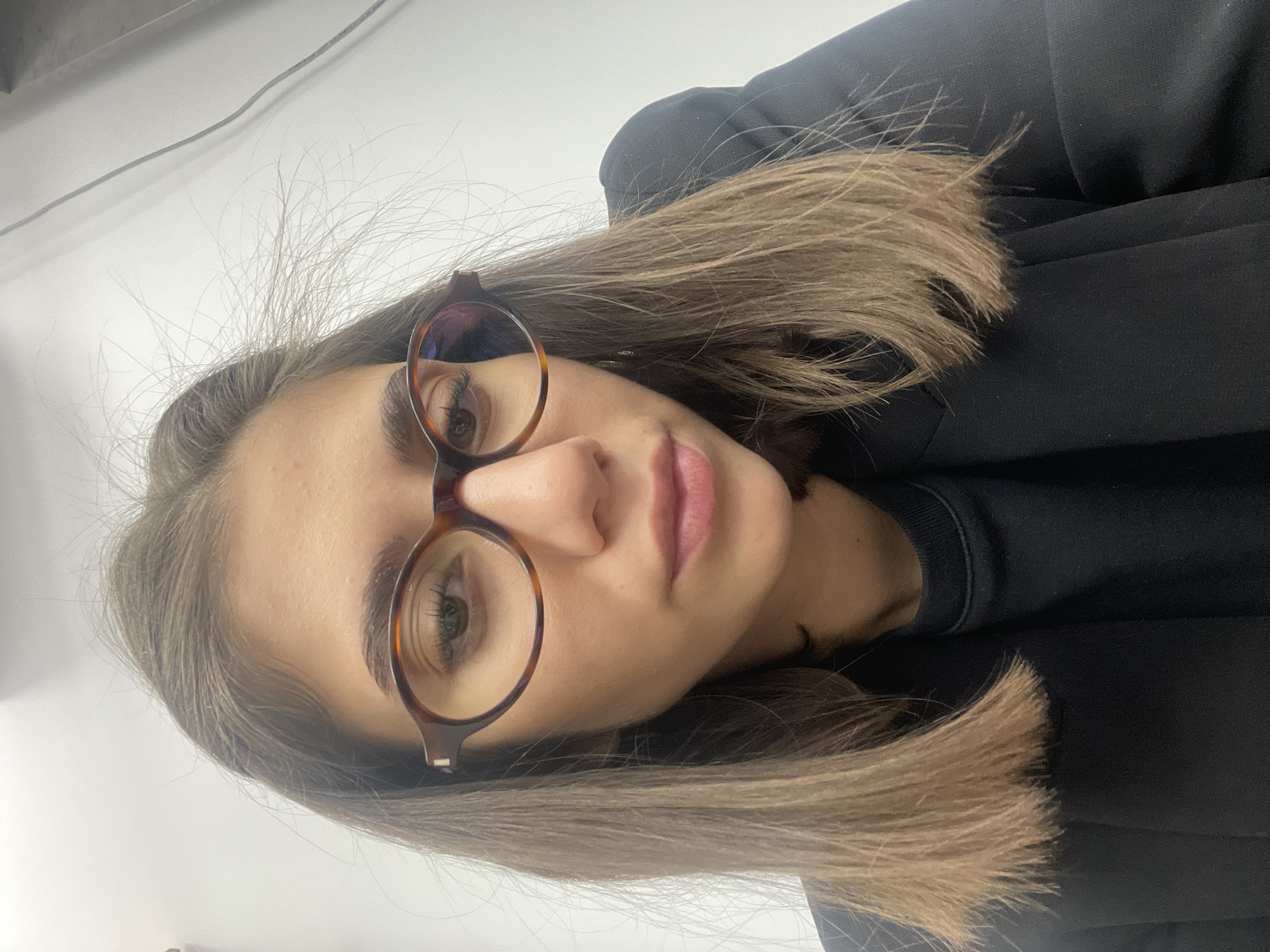 A close-up, portrait-style photograph, rotated 90 degrees clockwise, features a young woman looking directly at the camera. She has straight, light brown hair that reaches her shoulders and is parted to her left. Her eyes, appearing light in color (possibly green or hazel), are framed by tortoiseshell-rimmed glasses. Her eyebrows are dark and well-defined, and her eyelashes are prominent. Her lips are slightly parted, revealing a neutral expression. She is wearing a dark-colored, likely black, top or jacket. The background is a plain, light-colored wall, possibly white or light grey, with a thin, dark wire visible in the upper-left corner of the frame. The setting appears to be indoors. There are no discernible location cues within the image itself to confirm the provided context of Sofia, Bulgaria. No text or specific time of day indications are present. The woman's overall demeanor appears calm and composed.