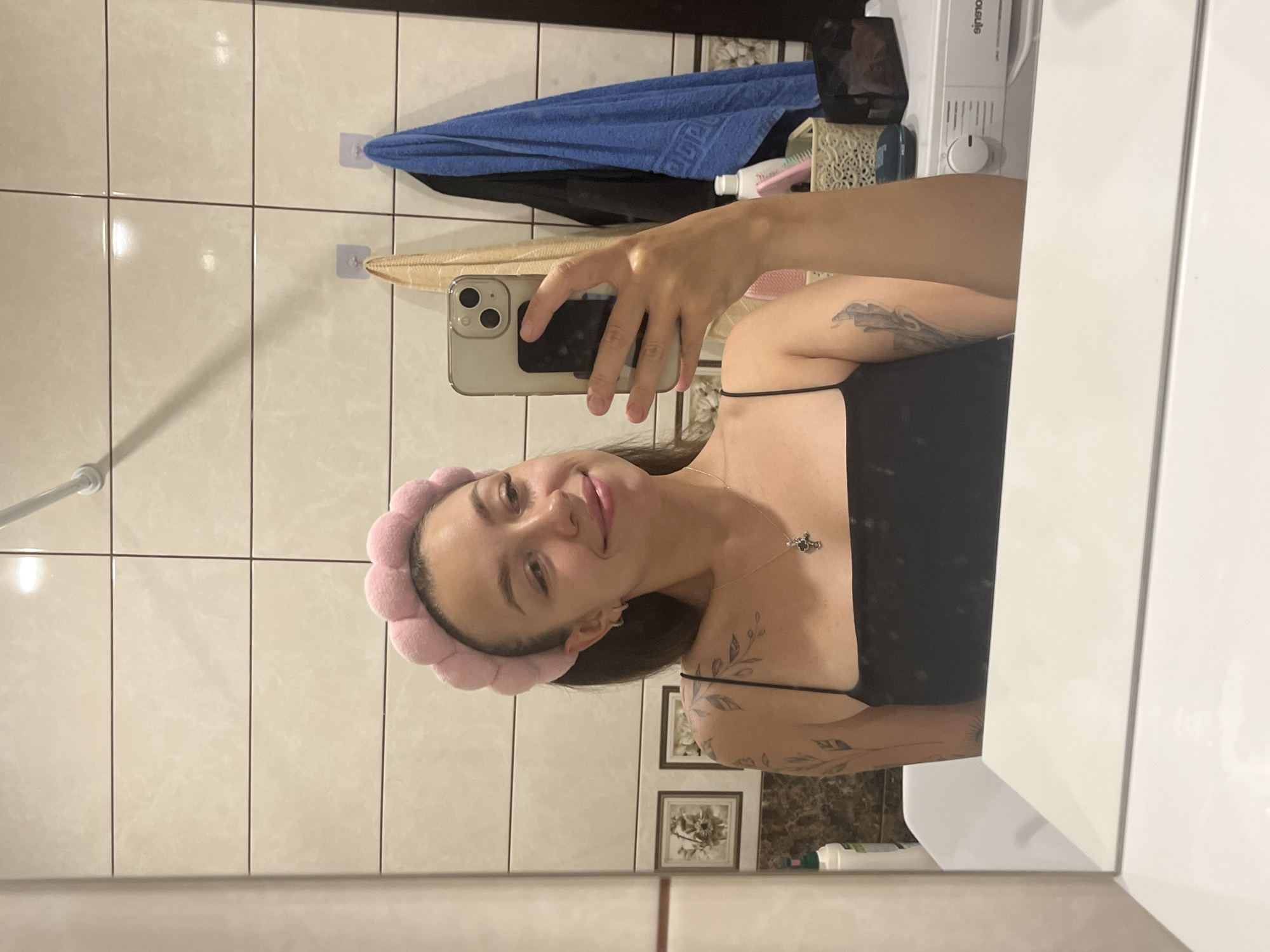 A young woman with a pink fuzzy headband and a black camisole is taking a mirror selfie in a bathroom. She is looking at her phone, which she is holding in her right hand. She has tattoos on her left arm and shoulder. In the background, there are beige tiles on the wall, a blue towel hanging, and a washing machine. The image appears to be taken indoors in a domestic setting.