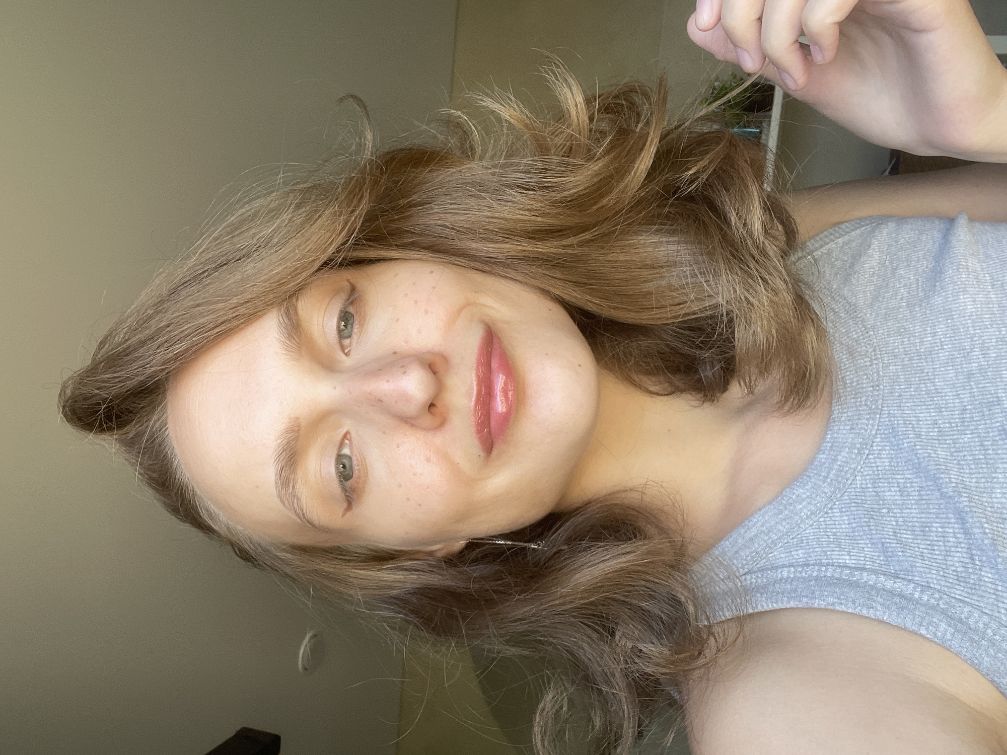 A medium shot depicts a woman with light brown, wavy hair and fair skin. She has freckles across her nose and cheeks. Her eyes are light-colored, and her lips are glossy. She is wearing a grey, sleeveless top. Her right hand is visible, touching a strand of her hair. The background is a plain, light-colored wall. It is impossible to determine if this image was taken in Подшивалово, Russia.
