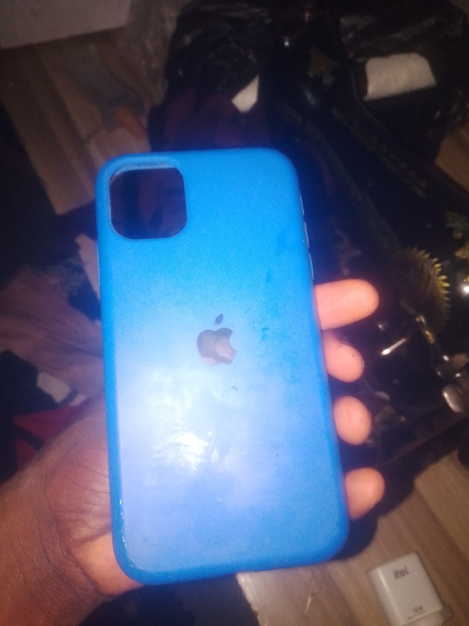 A hand is holding a bright blue phone case, which has an Apple logo embossed on it. The phone case is the primary subject of the image. The background is dark and out of focus, but appears to be indoors with a wooden surface in the foreground and some blurry objects in the background, possibly a sewing machine. A small white object with the 'itel' logo is visible in the lower right corner. The overall lighting is dim, suggesting the photo was taken in low-light conditions. There is no discernible indication of the location being Jauro Yinu, Nigeria.