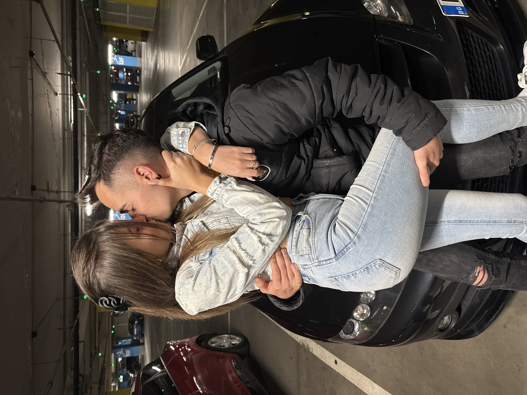 In an indoor parking garage in Sofia, Bulgaria, a young couple is intimately embracing and kissing on the hood of a black car. The man has short, dark hair with a defined undercut and wears a black padded jacket and dark jeans. The woman has long brown hair and wears a light blue denim jacket and light blue jeans. The car's license plate is visible, indicating Bulgarian origin. The background reveals multiple levels of the parking structure with signs and parked vehicles.