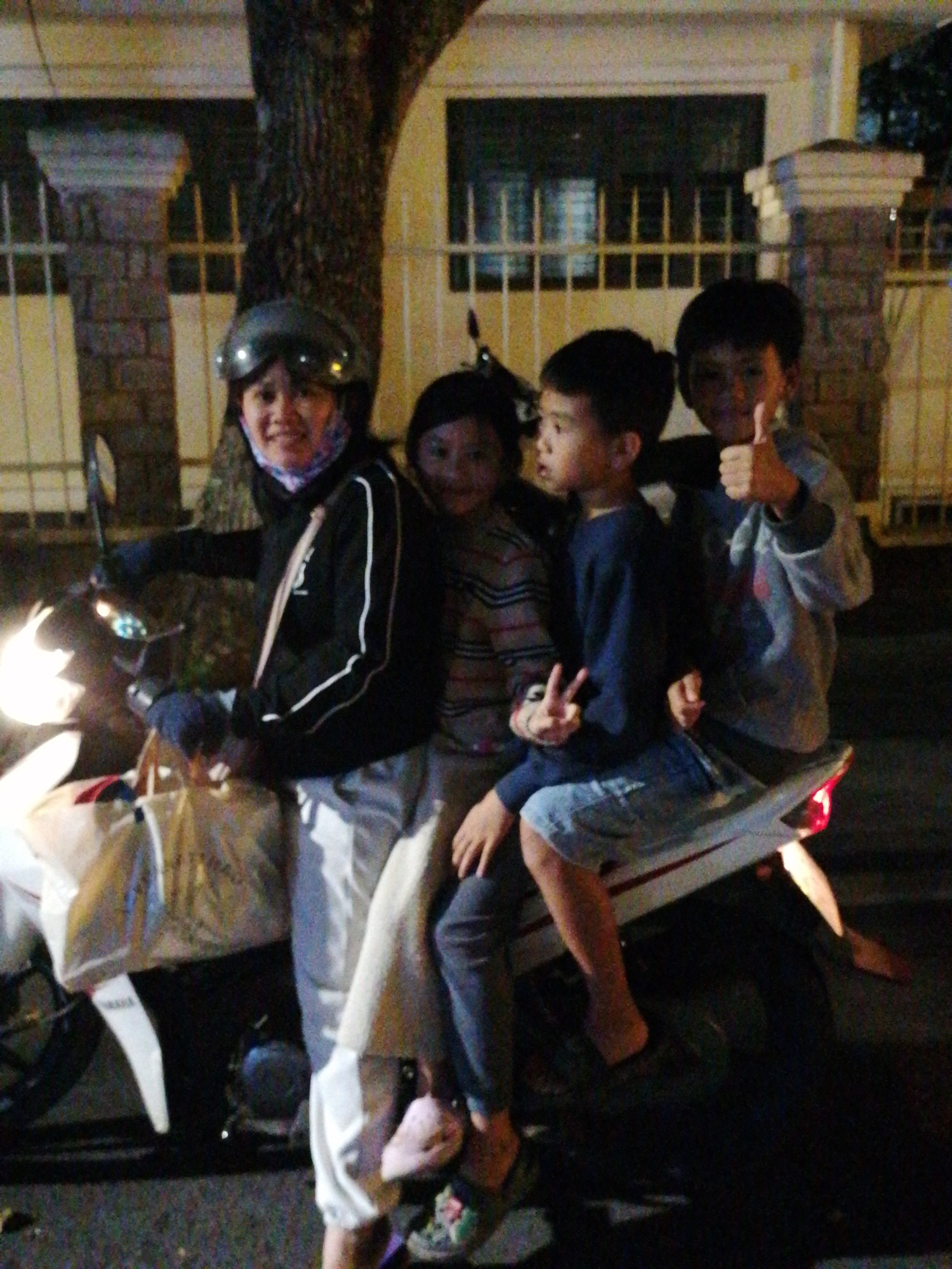 Four individuals, including an adult woman and three boys, are seated on a motorbike at night. The woman, wearing a helmet and a black jacket with white stripes, is operating the vehicle. The three boys are positioned behind her. The two younger boys are seated in front of the elder boy. One of the younger boys is holding his hand up in a peace sign. They are carrying several bags on the front of the motorbike. The background suggests an urban environment, possibly in Da Lat, Vietnam, with a fence and a tree visible. The lighting indicates it is nighttime, with the motorbike's headlight illuminated.