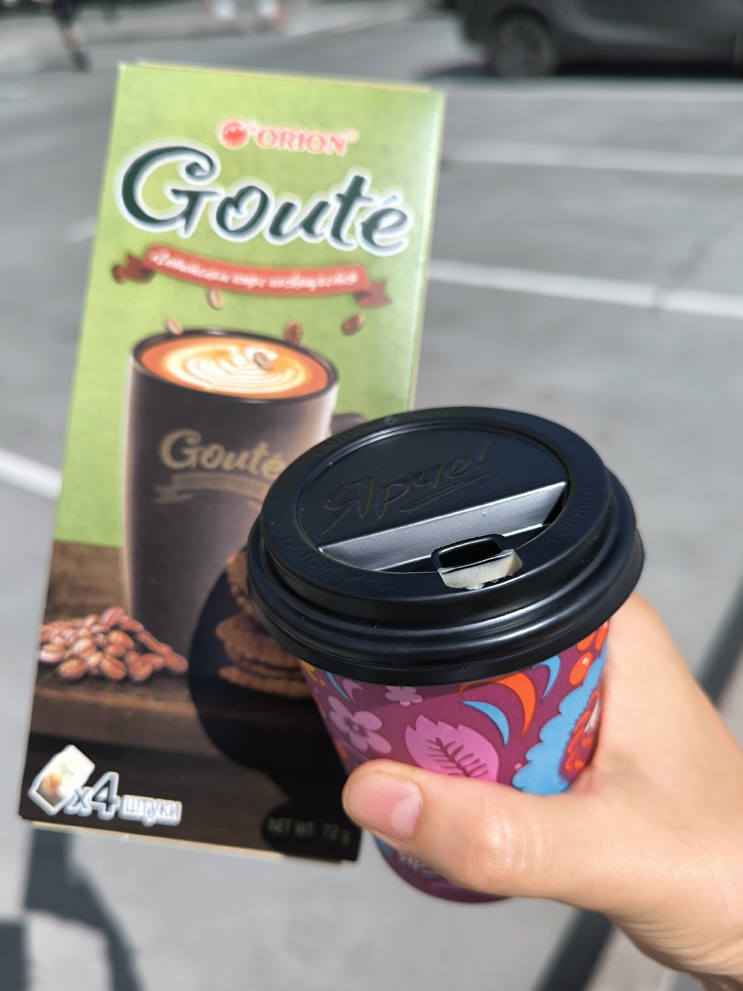 An outdoor, eye-level medium shot shows a person's right hand holding a paper coffee cup with a black lid and a colorful floral design. The cup is in the foreground, and a box of "Goute" coffee mix, with an image of a coffee cup with latte art and coffee beans, is behind it. The background is blurred, showing a paved surface with white lines, suggesting a parking lot or street in Novosibirsk, Russia. The overall lighting is bright and sunny.