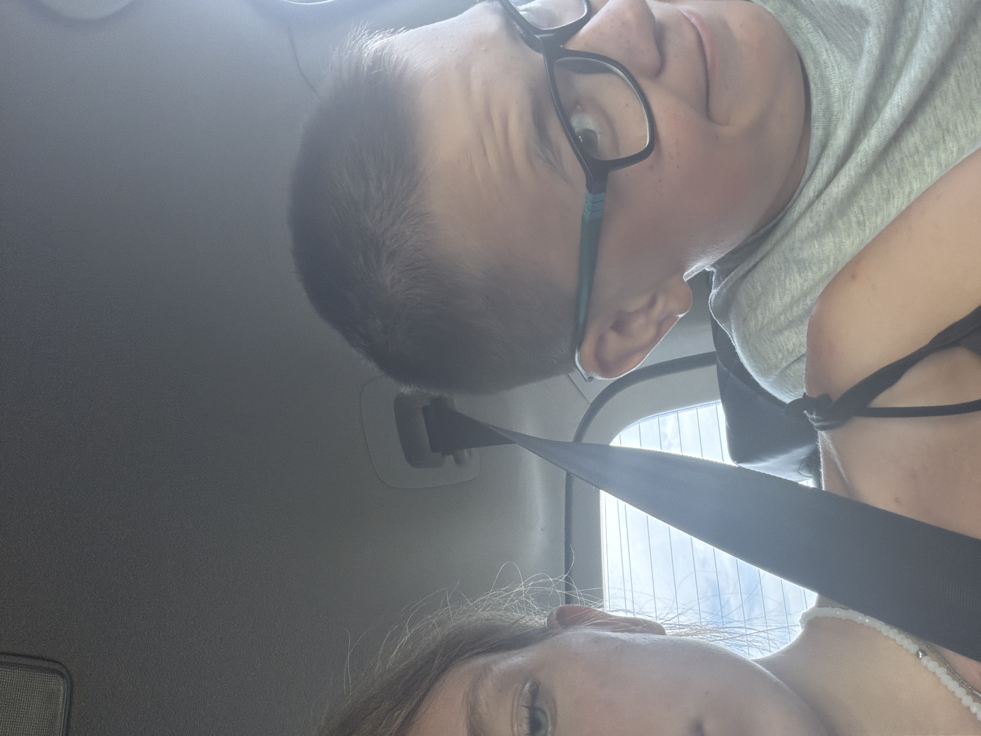 In a vehicle, a young person with blond hair and wearing a necklace is on the left, looking slightly off-camera with their left eye visible. To the right, a young person with short brown hair, wearing glasses and a light gray t-shirt, is looking towards the camera with a slight smile. A black seatbelt is visible in the foreground, diagonally crossing the image. The background shows the interior of a car, including the headliner and a window with a view of the sky and clouds, suggesting the vehicle is in motion. The lighting appears to be natural daylight coming from outside the car. The location is Teneevo, Russia.
