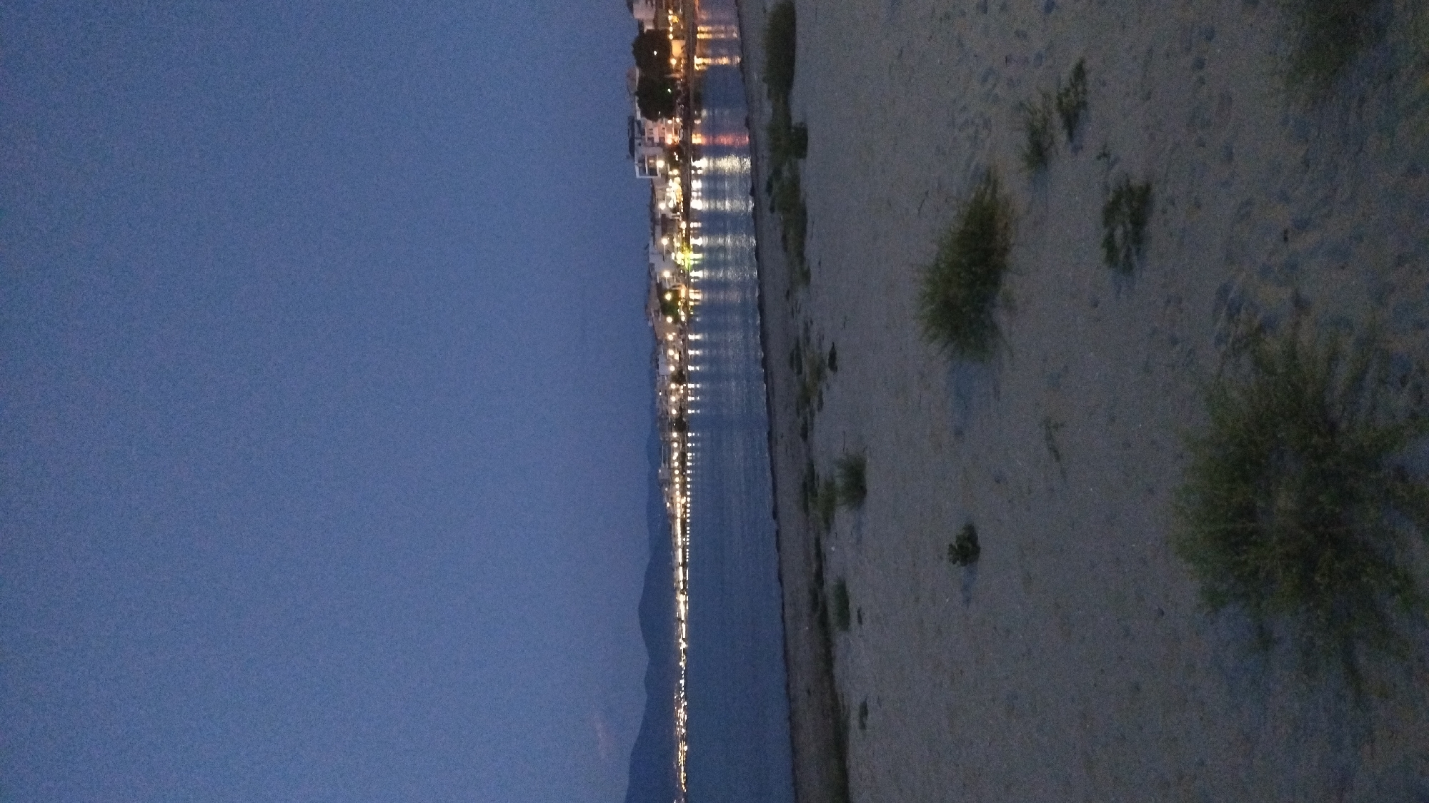 The image depicts a nighttime view of the coastline in Neoi Epivates, Greece. In the foreground, a sandy beach is visible with sparse vegetation. Beyond the beach, the calm sea reflects the lights of a town situated along the shoreline. The town appears to have low-rise buildings and is illuminated by numerous streetlights and building lights, creating a warm glow on the water. In the distance, dark hills or mountains form a backdrop against the twilight sky. The overall atmosphere is serene and peaceful, characteristic of a coastal town at dusk or night.