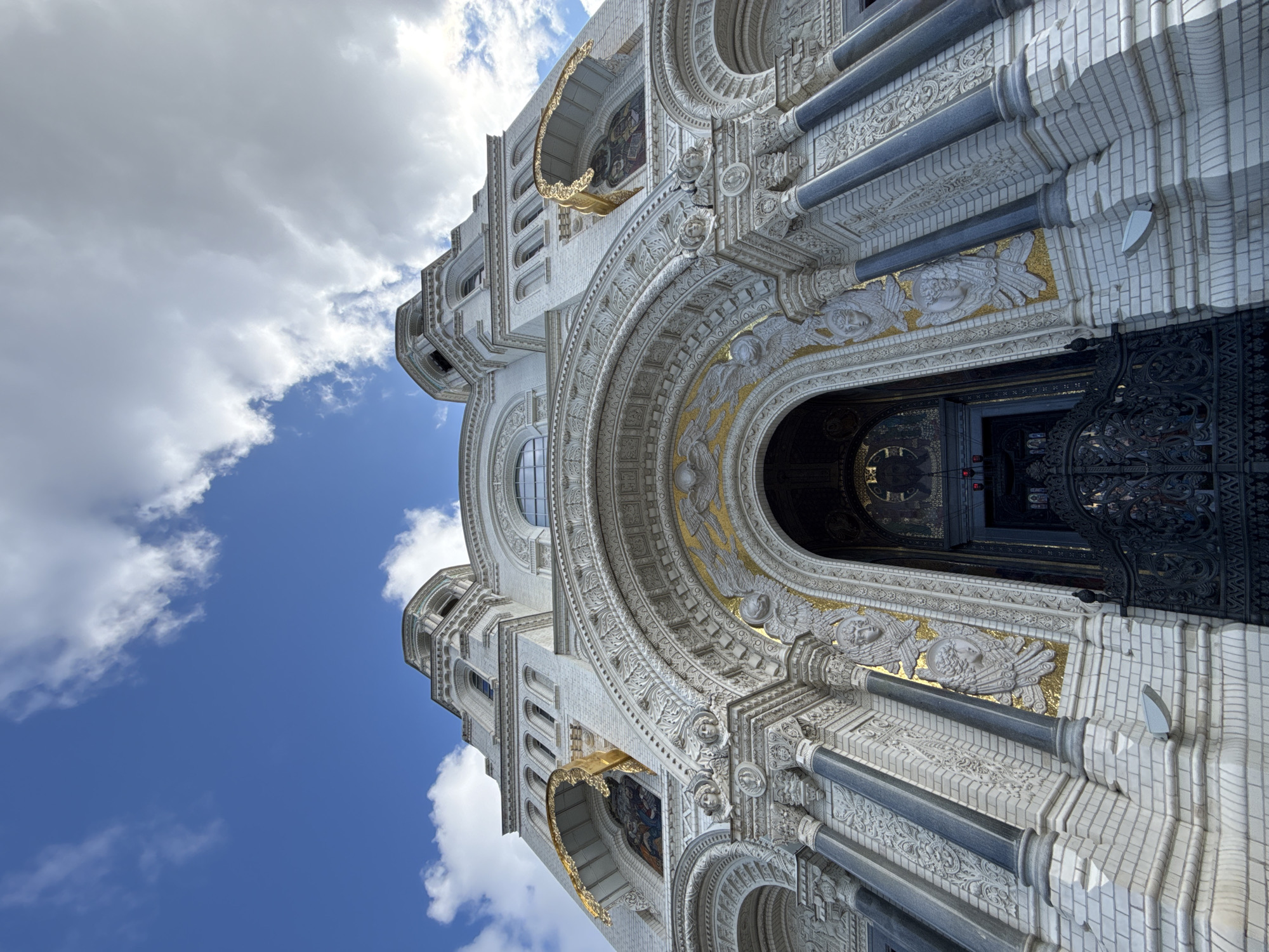 The image shows a low-angle, close-up view of the facade of the Naval Cathedral of Saint Nicholas in Kronstadt, Russia. The cathedral is a large, white, brick building with intricate architectural details, including arched doorways, ornate carvings, and gilded mosaic elements depicting angels. Several columns and turrets are visible, along with a large black metal gate in front of the main entrance. The sky above is a mix of blue with scattered white and gray clouds. The lighting suggests it is daytime, with some shadows cast on the building.