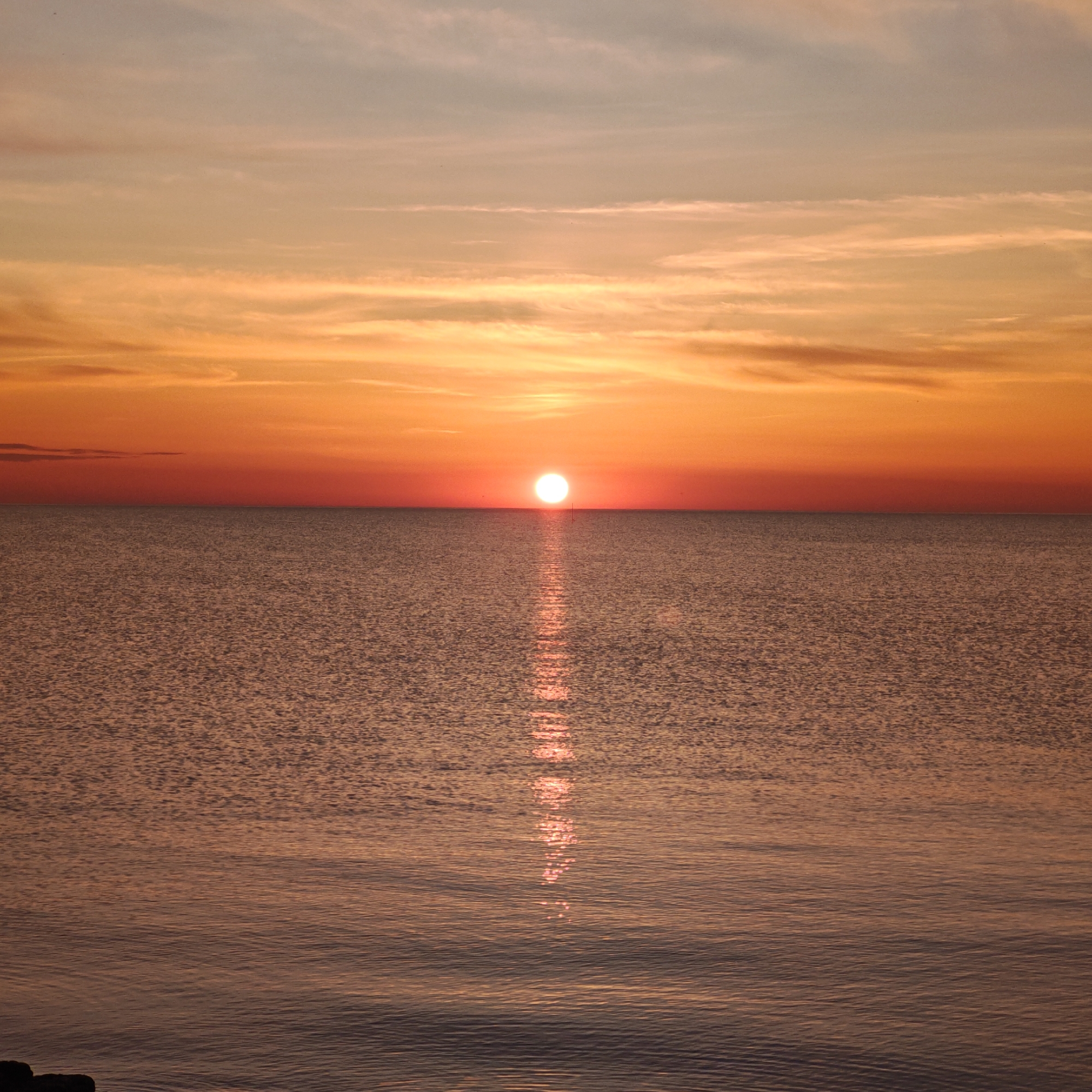 The image depicts a sunset over the sea. The sun is positioned on the horizon, casting a bright reflection across the water towards the viewer. The sky exhibits varying hues of orange and yellow, with some cloud formations visible. The sea appears calm, with small ripples visible on the surface. There are no people or objects visible on the water or the shore. The location is the city of городское поселение Щелкино, Ukraine.