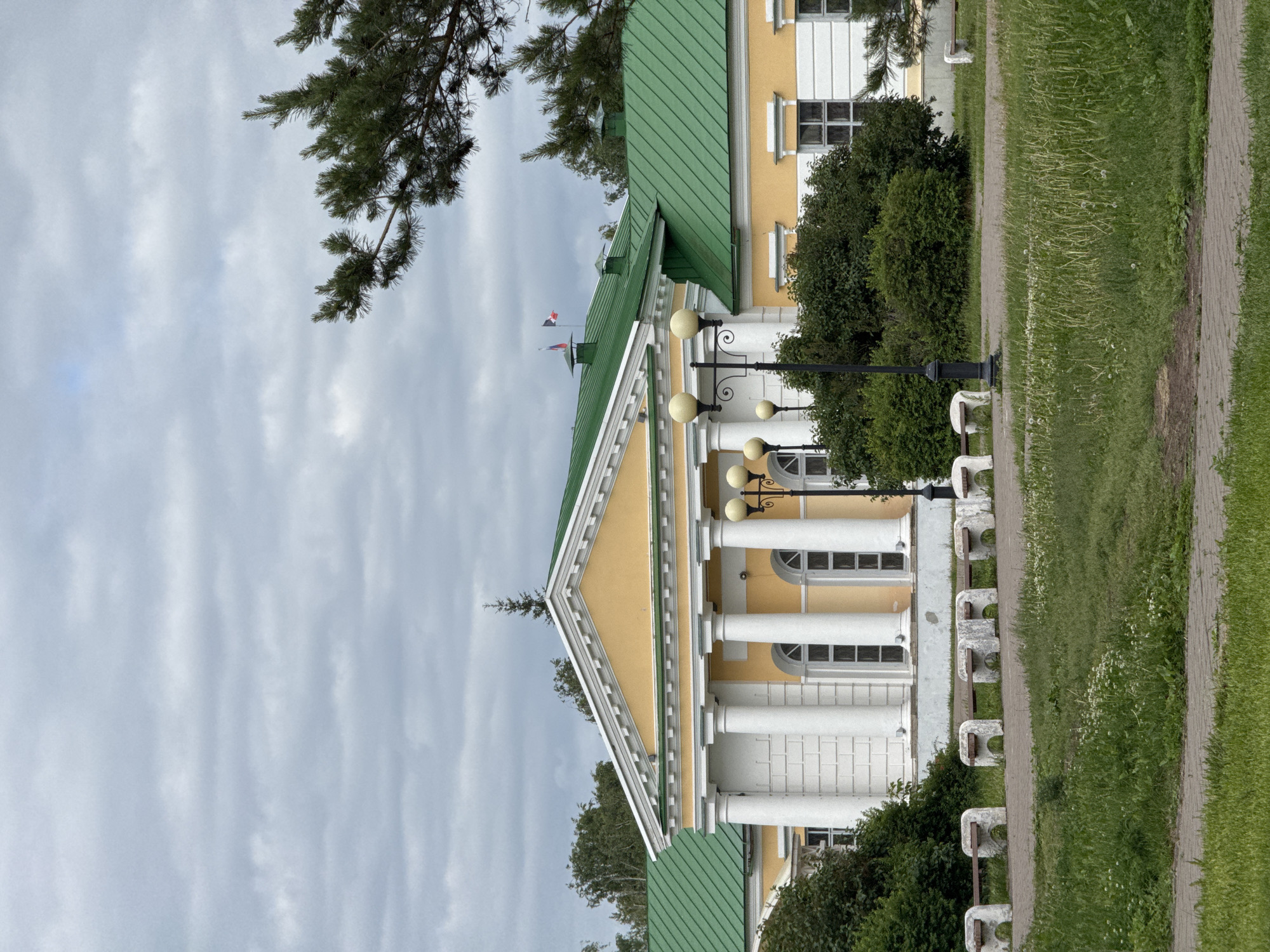 The image shows a neoclassical building with a yellow facade and a green roof, featuring a portico with white columns. The building is situated in a park-like setting with manicured lawns, paved walkways, and several concrete benches with wooden seats. A tall lamppost with multiple light fixtures stands in the foreground. Trees, including a pine tree, are visible around the building. Two flags, one Russian and one with a tricolor pattern, are flying from the roof of the building. The sky is overcast with grey clouds. The building is identified as the Museum of Fine Arts in Izhevsk, Russia.