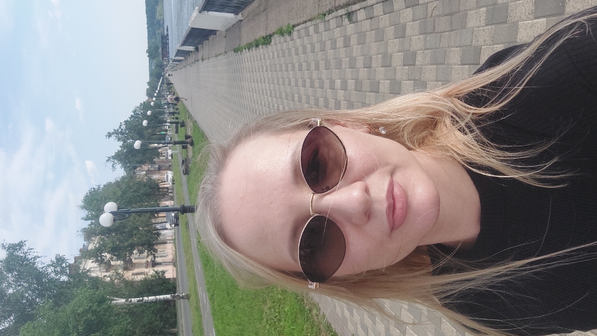 A young woman with blonde hair and sunglasses is taking a selfie outdoors on a paved walkway. She is wearing a black top. In the background, a tree-lined street with buildings and cars is visible, as is a body of water. The scene appears to be a promenade or embankment in Votkinsk, Russia, a city located on the Kama River.