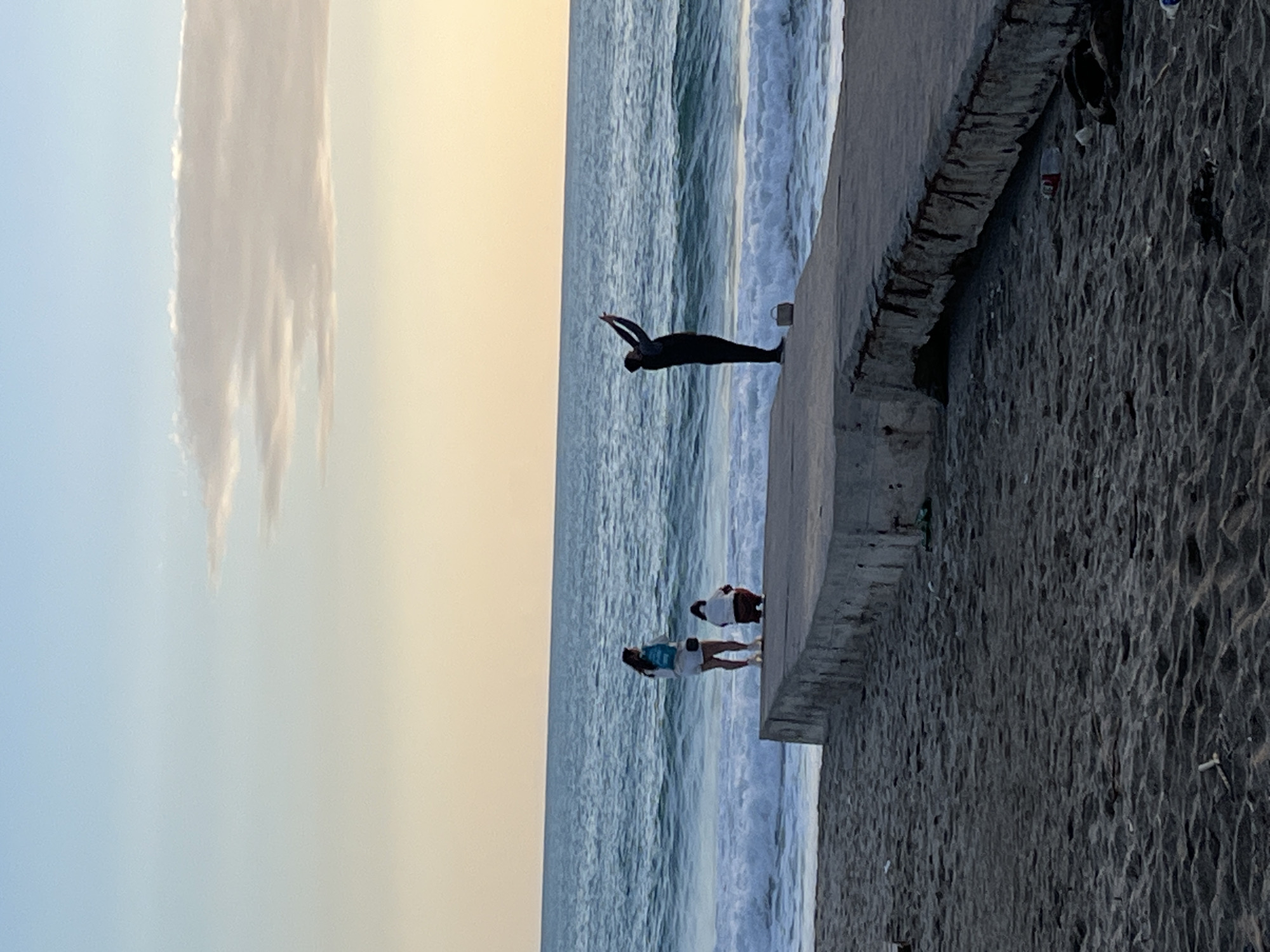 The image depicts a beach scene in Alanya, Turkey. The camera is positioned on the shore looking out at the sea. The beach is dark sand. There is a stone pier on the right-hand side of the frame. The sea is visible with waves breaking towards the shore. In the distance, a person in a dark, tight-fitting outfit is diving into the water from the pier. Further along the pier, near the shoreline, two people are standing at the edge, looking out to sea. The sky is pale, with a vertical band of clouds on the left side.