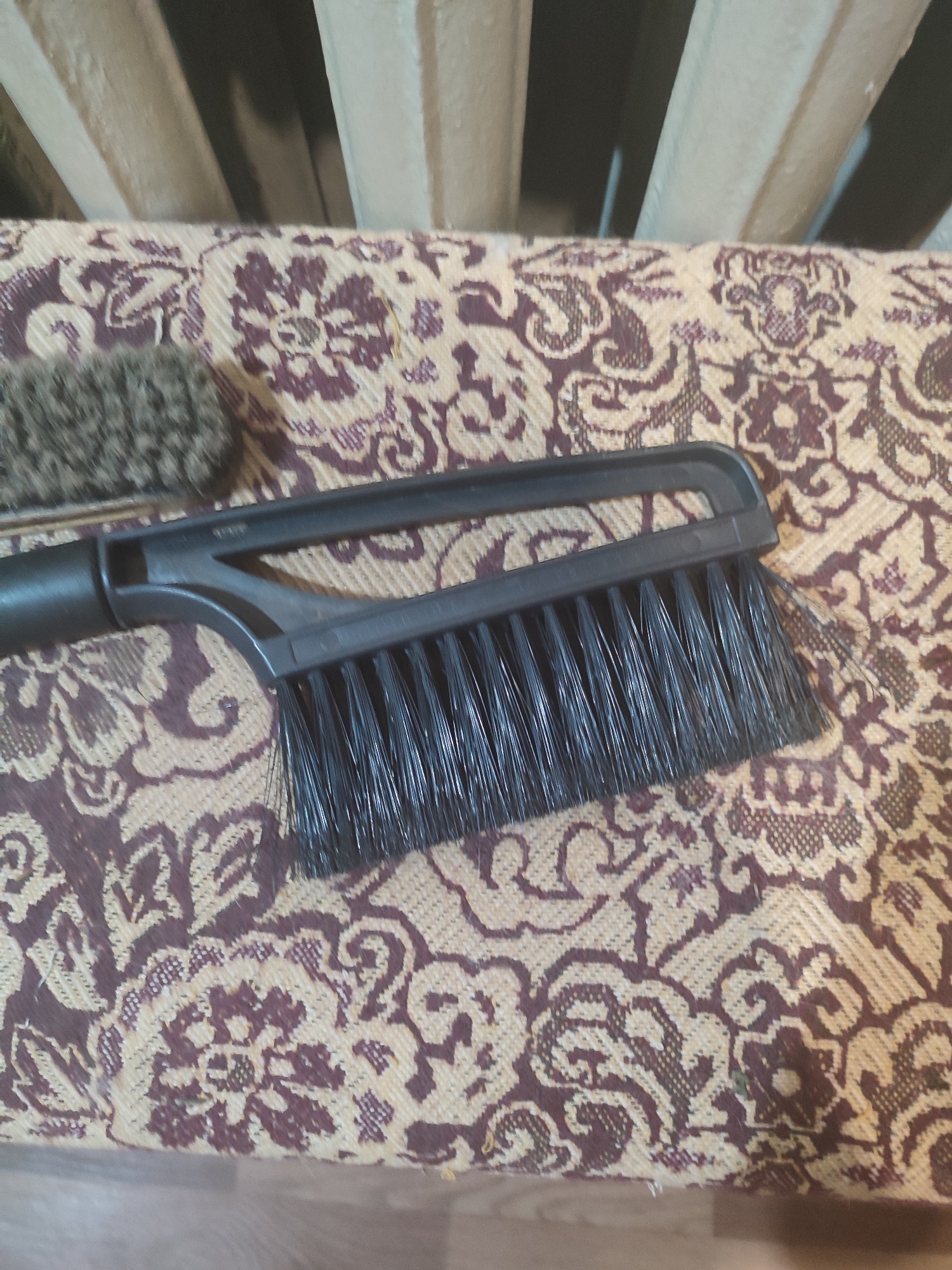 The image shows a black handled brush with stiff bristles resting on a patterned fabric, likely a chair or sofa. The fabric has a repeating floral design in shades of beige and dark red. In the background, a section of a white radiator is visible. To the left of the main brush, a portion of another brush, appearing to be made of a coarser material, is also visible. The lighting suggests an indoor environment. The location is identified as Narowlya, Belarus.