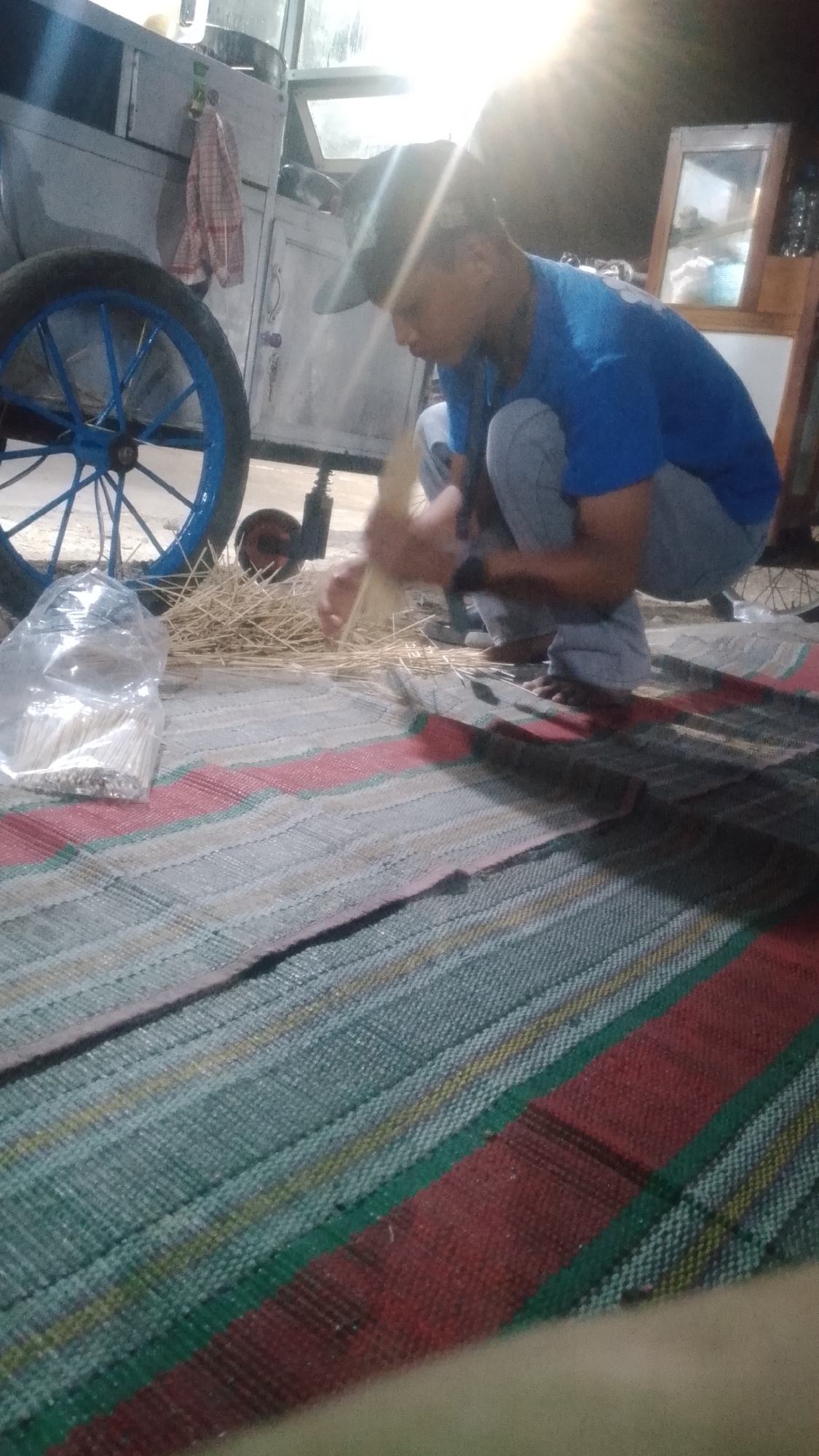 A young male, wearing a blue t-shirt and gray pants, crouches on a woven mat in what appears to be an outdoor setting at night. He is focused on processing a bundle of dried grass or reeds. A large blue-rimmed wheel from a cart is visible to his left, suggesting a roadside or market stall environment. A plastic bag filled with wooden skewers is on the mat in front of him. The background shows a cart with a towel hanging from it and a brightly lit window, possibly indicating a food stall or workshop. The overall lighting suggests artificial light sources are in use.