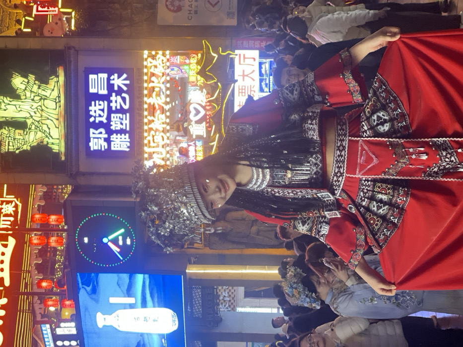 A young East Asian woman, centrally positioned and facing slightly left with a subtle smile, is featured outdoors at night in Yuzhong District, Chongqing, China, likely within the Hongyadong Commercial Street area given the architectural and commercial context. She wears a vibrant red, two-piece traditional costume adorned with intricate silver and black patterns, extensive silver metallic embellishments, and fringe detailing. A large, ornate silver-colored headdress with leaf-like elements crowns her head, and a prominent silver necklace is visible. White glitter or makeup is applied near her eyes. She holds out the bottom edges of her skirt with both hands.
The background is a densely illuminated commercial street at night. Above and to the left of the woman, a large digital billboard displays a white bottle and a green-rimmed circular graphic. To her right, a blue neon sign reads "郭选昌 雕塑艺术" (Guo Xuanzhang Sculpture Art), below which is a prominent yellow and red neon sign for "九宫格老火锅" (Jiugongge Old Hotpot). Further signs, including a partial "CHAGEE" logo and "售票大厅" (Ticket Hall), are visible. Red traditional lanterns hang in the upper left. A dense crowd of people occupies the background; one woman to the left wears light blue traditional attire with a floral headpiece and is looking at a mobile phone. The scene is bustling with pedestrian traffic and vibrant commercial lighting.