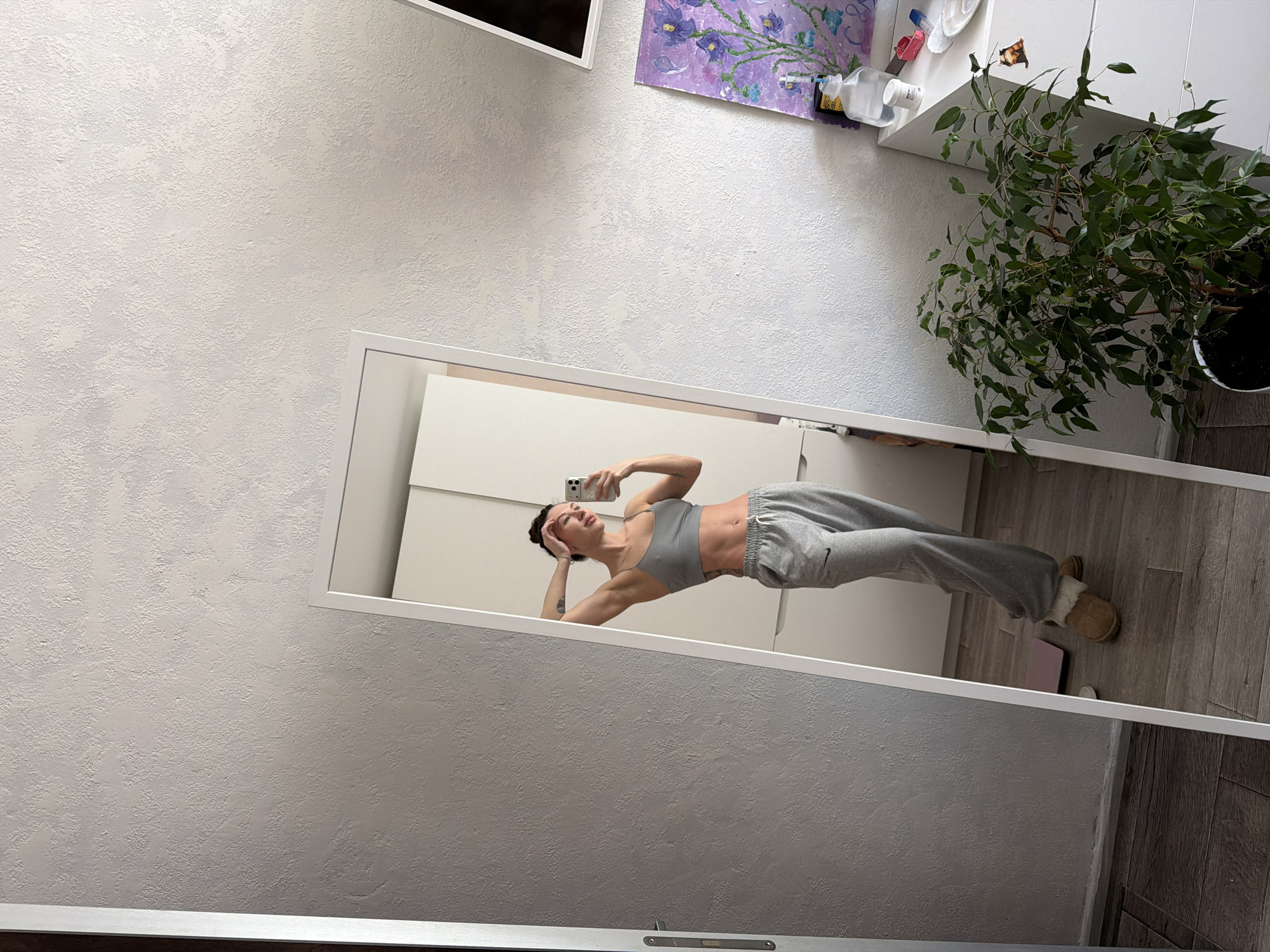A woman in gray workout clothes and slippers is taking a selfie in a full-length mirror. The mirror is leaning against a white wall in a room with gray, textured walls. A painting and other objects are on a shelf near the top of the frame. A large houseplant is visible on the right side of the image. The location is Perm, Russia.