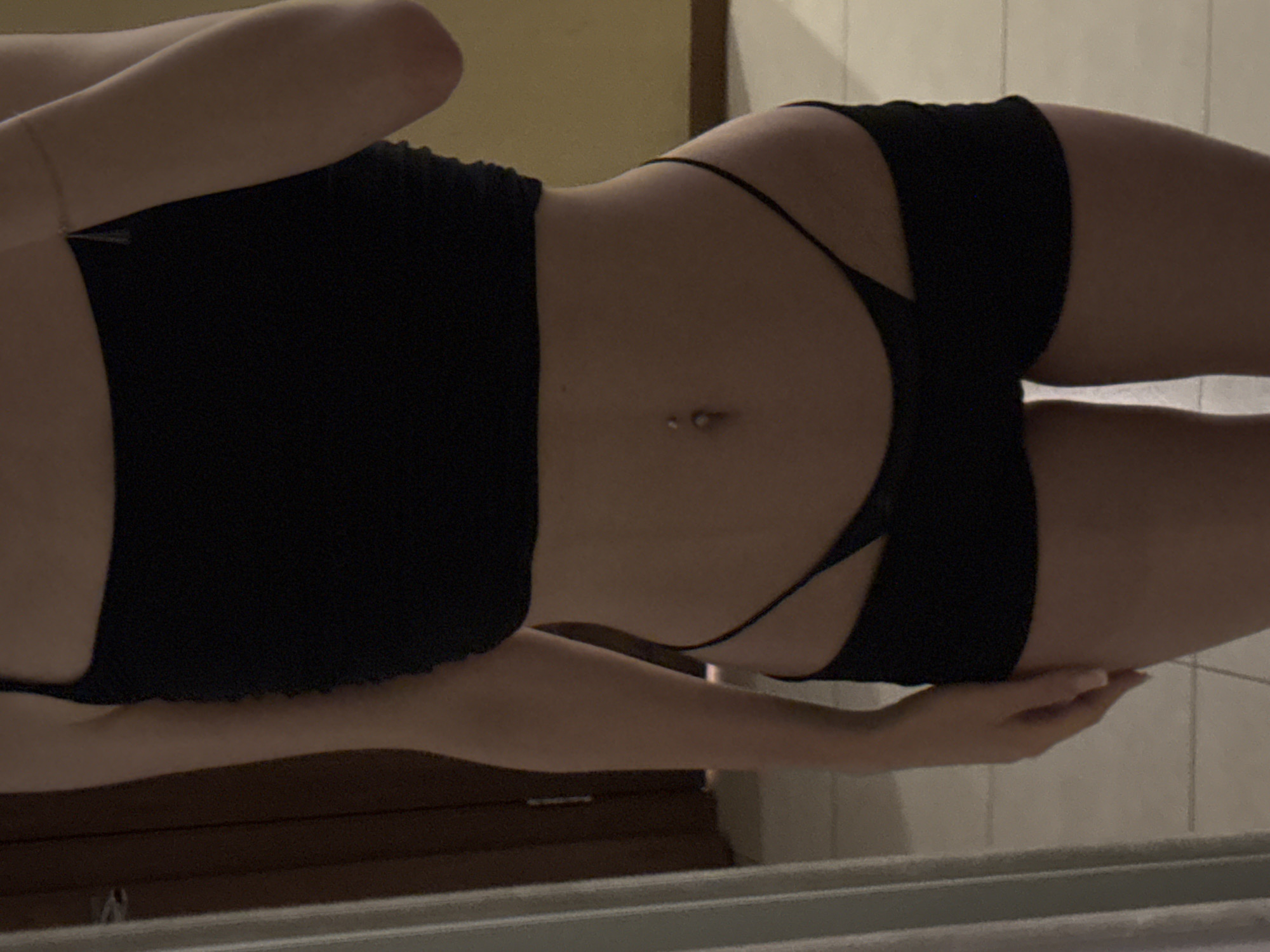The image shows a person's midsection, with a focus on their stomach and hips. They are wearing black underwear that appears to be a thong and a matching top. The background is blurred, but it seems to be an indoor setting with tiled walls and some wooden furniture visible. The lighting is dim, suggesting it might be nighttime or a poorly lit room. There is a belly button piercing visible. The person's hand is resting on their hip. The image is oriented vertically.