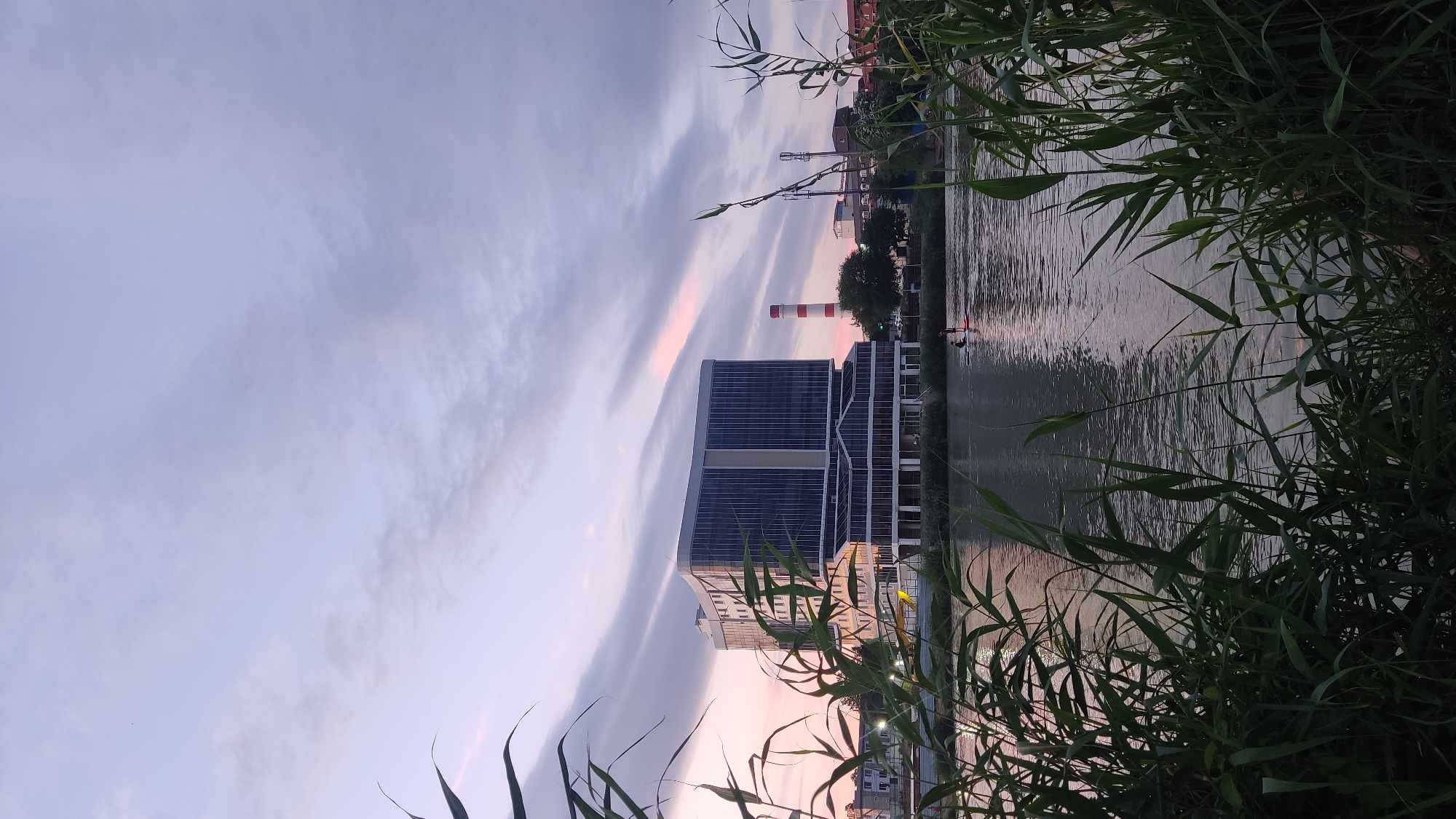 The image depicts a picturesque sunset over the Kuban River in Krasnodar, Russia. In the foreground, lush green reeds frame the scene, with their leaves gently swaying in the breeze. The river's surface reflects the soft hues of the twilight sky, creating a serene and tranquil atmosphere.
Dominating the mid-ground is a modern, multi-story building with a sleek, glass facade. Its clean lines and contemporary design stand in stark contrast to the natural beauty of the river and sky. Adjacent to the building, a tall, red-and-white striped chimney rises into the sky, hinting at industrial activity in the vicinity.
Further down the riverbank, other buildings and trees can be seen, suggesting a vibrant urban landscape merging with nature. Two individuals are visible on the water, engaged in what appears to be paddleboarding, adding a touch of human activity to the otherwise calm scene. The sky is adorned with wispy clouds, tinged with shades of pink and purple from the setting sun, casting a magical glow over the entire panorama.